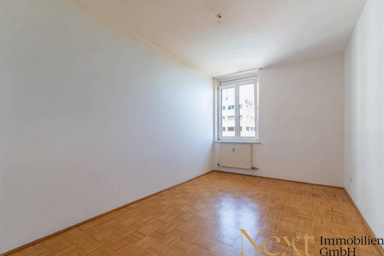 Leeres Schlafzimmer mit Parkettboden und einem Fenster, das auf ein gegenüberliegendes Gebäude blickt.