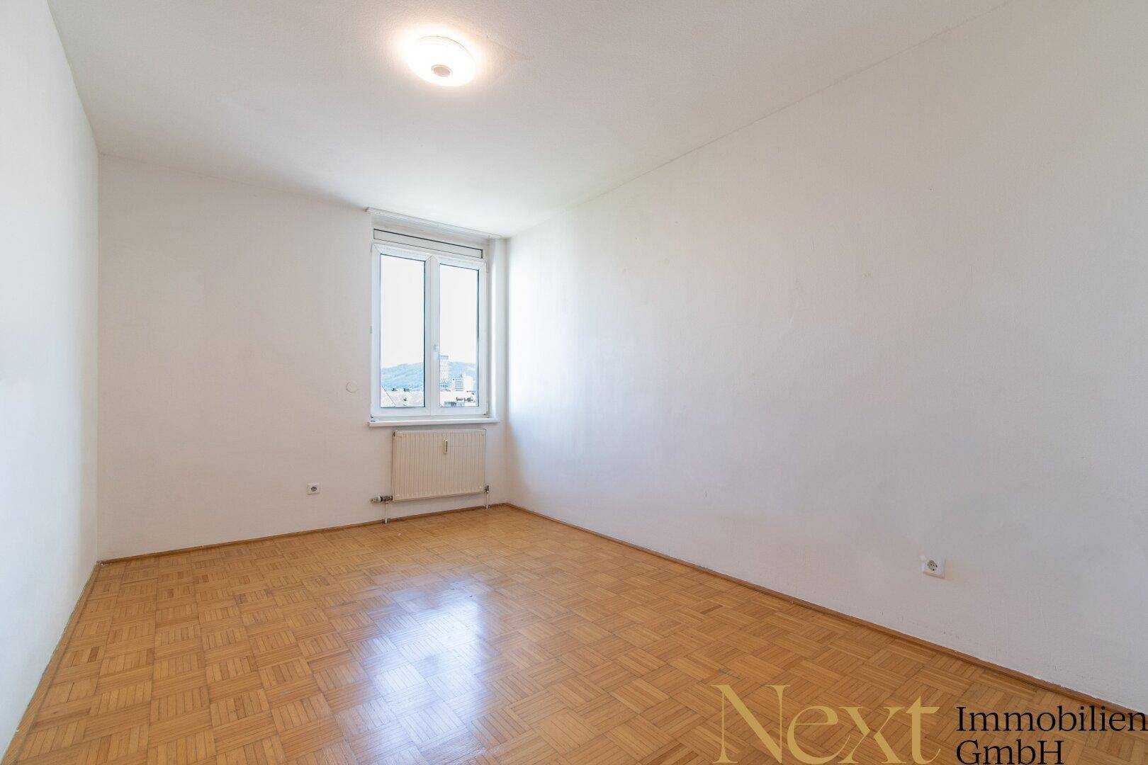 Helles, leeres Zimmer mit Parkettboden und einem Fenster, das einen weiten Ausblick ermöglicht.