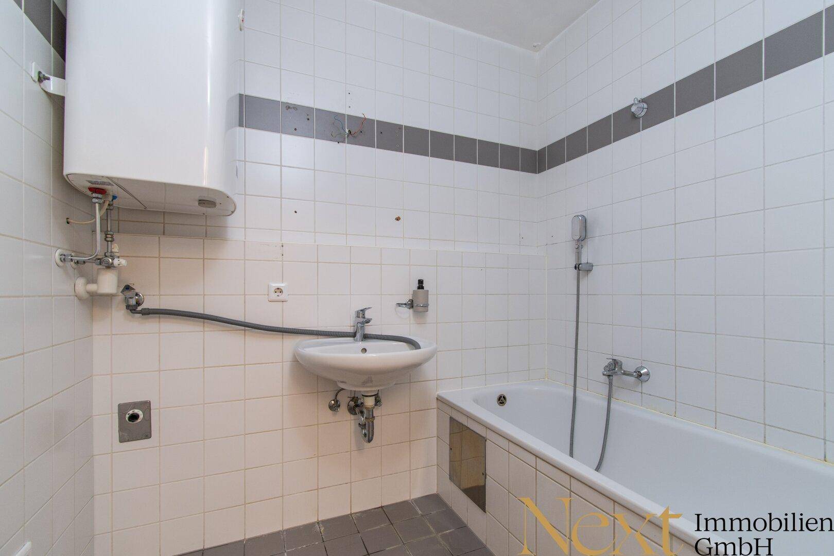 Einfaches Badezimmer mit weißen Fliesen, kleiner Badewanne, Waschbecken und Warmwasserboiler.