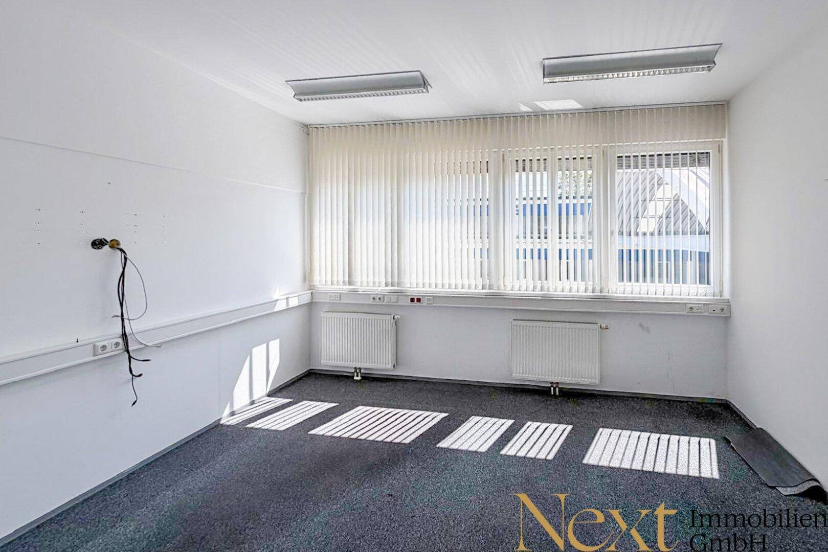 Leeres Büro mit grauem Teppichboden, weißen Wänden, Fenstern mit Jalousien und Heizkörpern.