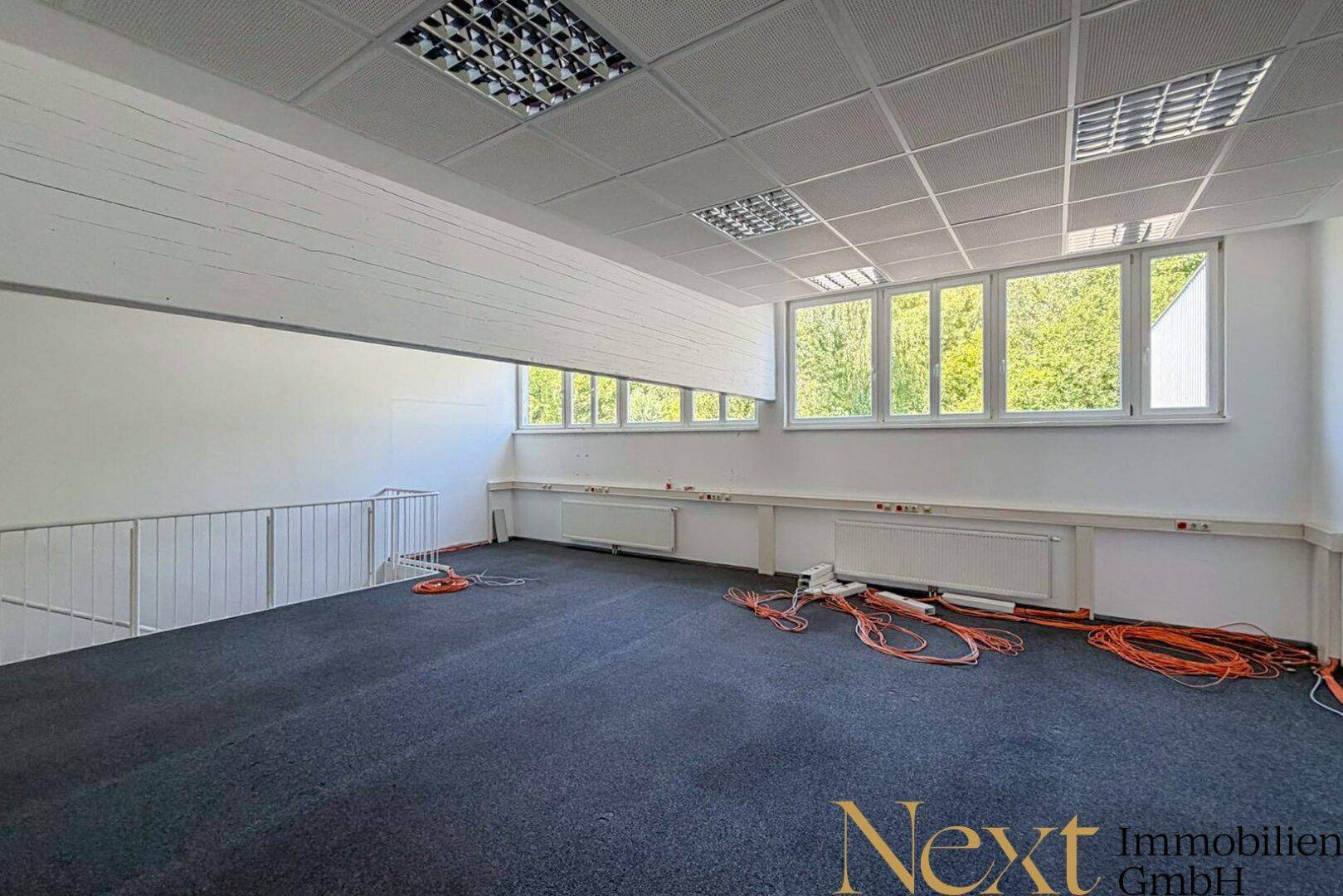Offener Bürobereich auf einer Empore mit Teppichboden, großen Fenstern und abgehängter Decke.
