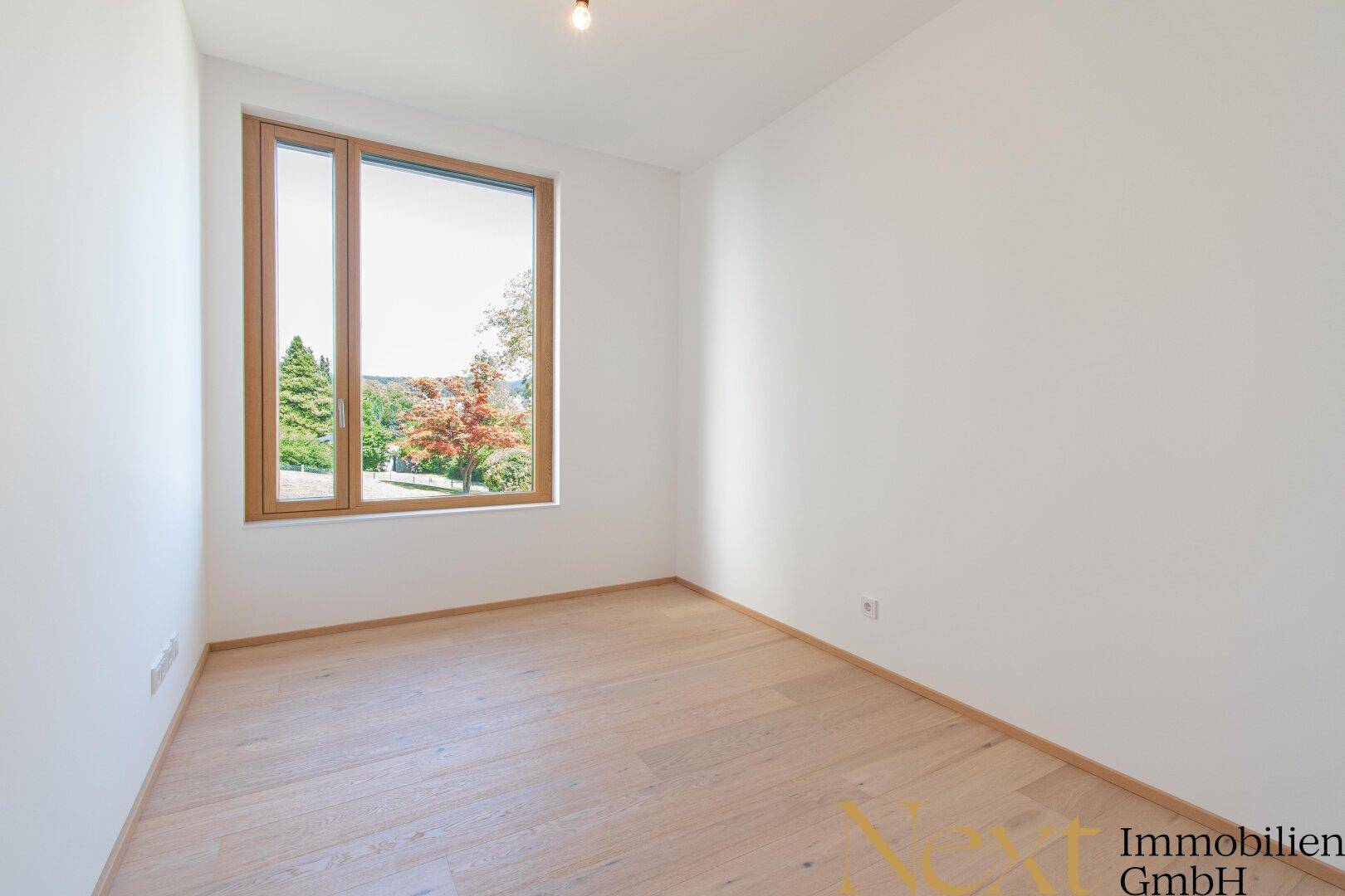 Leeres Schlafzimmer mit hellem Holzboden und großem Fenster mit Blick ins Grüne.