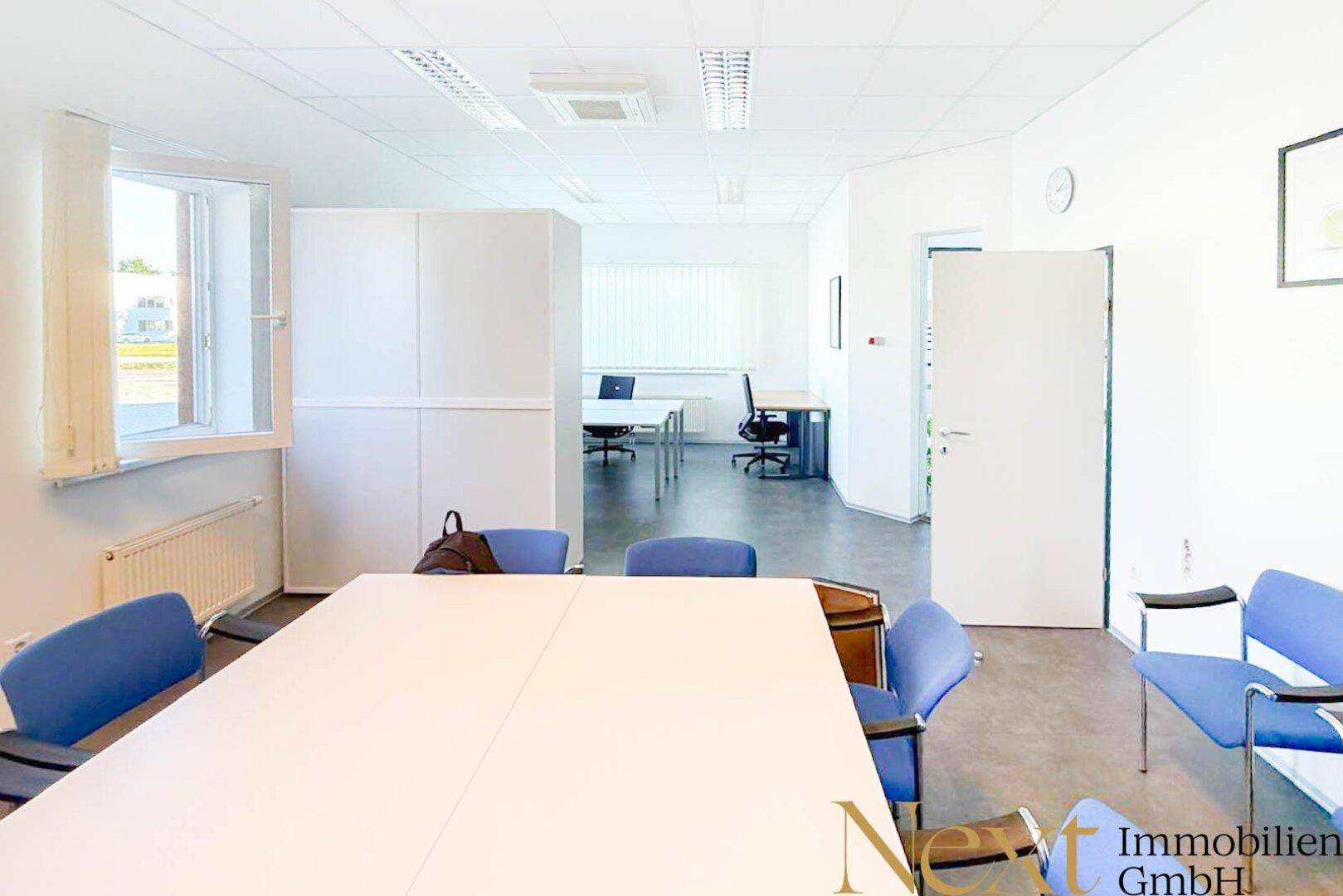 Heller Büroraum mit großem Konferenztisch und offenem Arbeitsbereich im Hintergrund, ausgestattet mit modernen Möbeln.