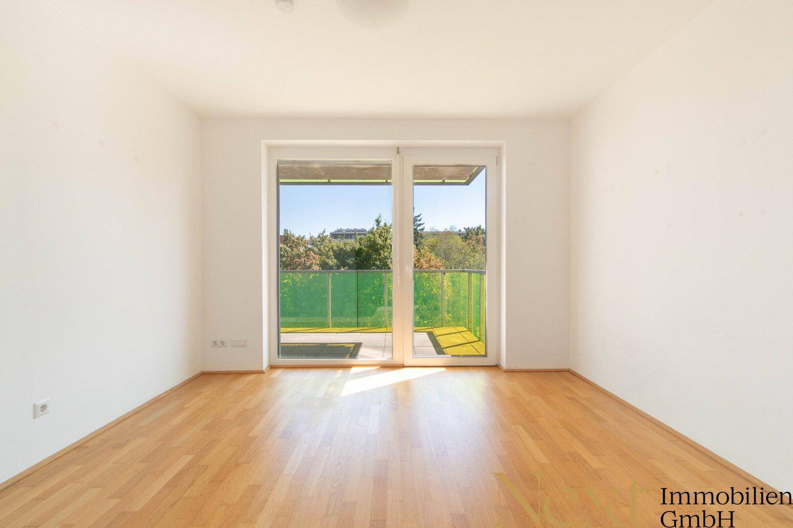 Leerer Raum mit Holzboden und großem Fenster, das Zugang zu einem Balkon mit grüner Brüstung bietet.
