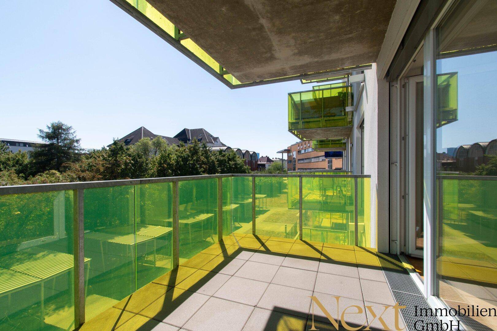 Geräumiger Balkon mit grünen Glasbrüstungen, Fliesenboden und Blick ins Grüne.