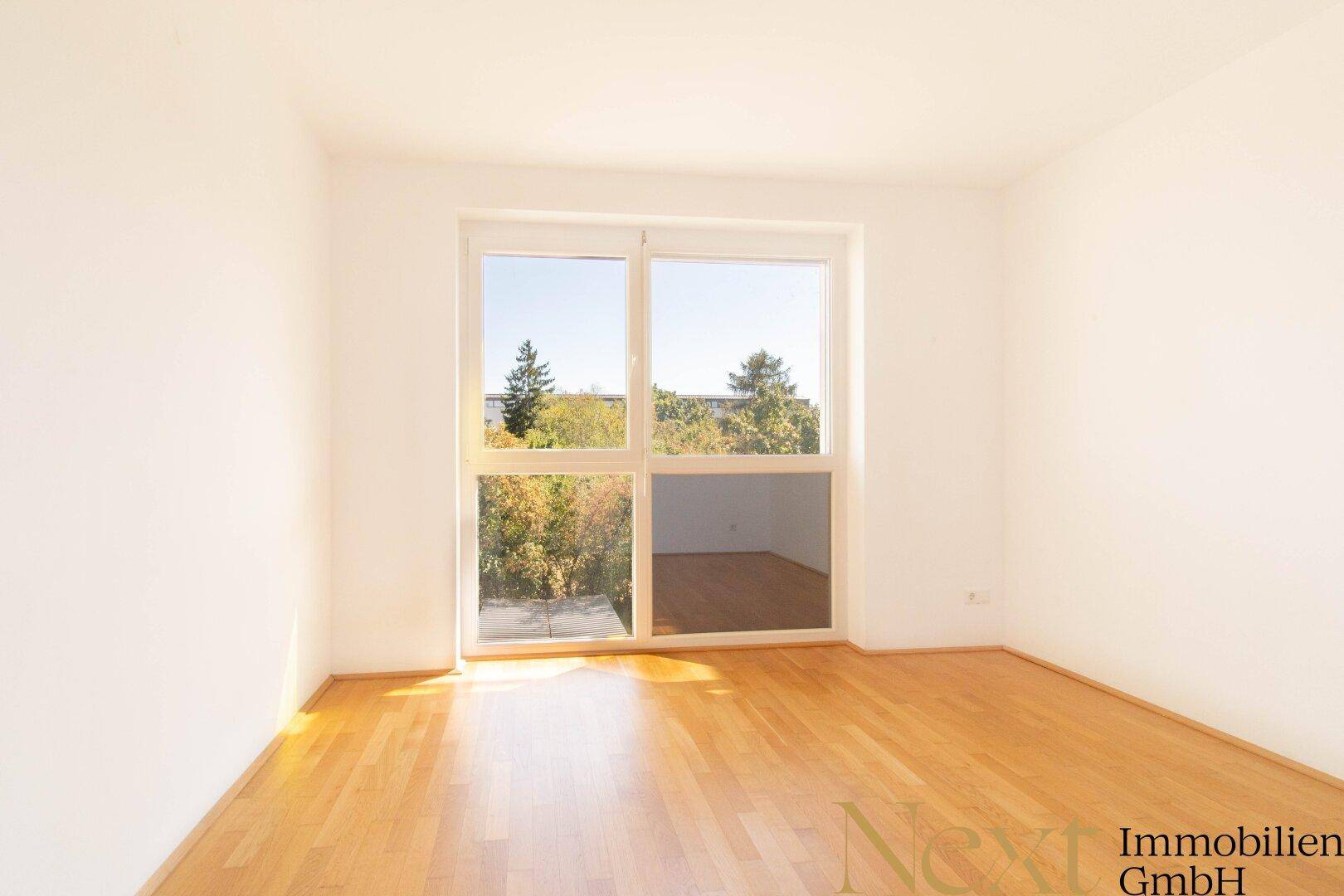 Heller, leerer Raum mit Holzboden und großem Fenster, das viel Tageslicht hereinlässt.