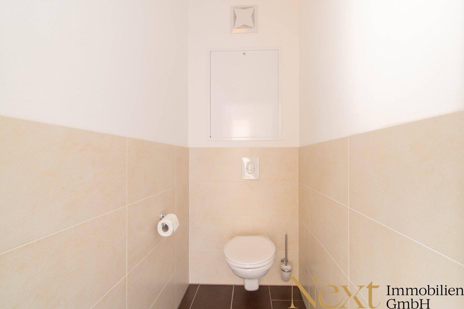 Separates WC mit Wandtoilette, beigen Fliesen und dunklem Boden.