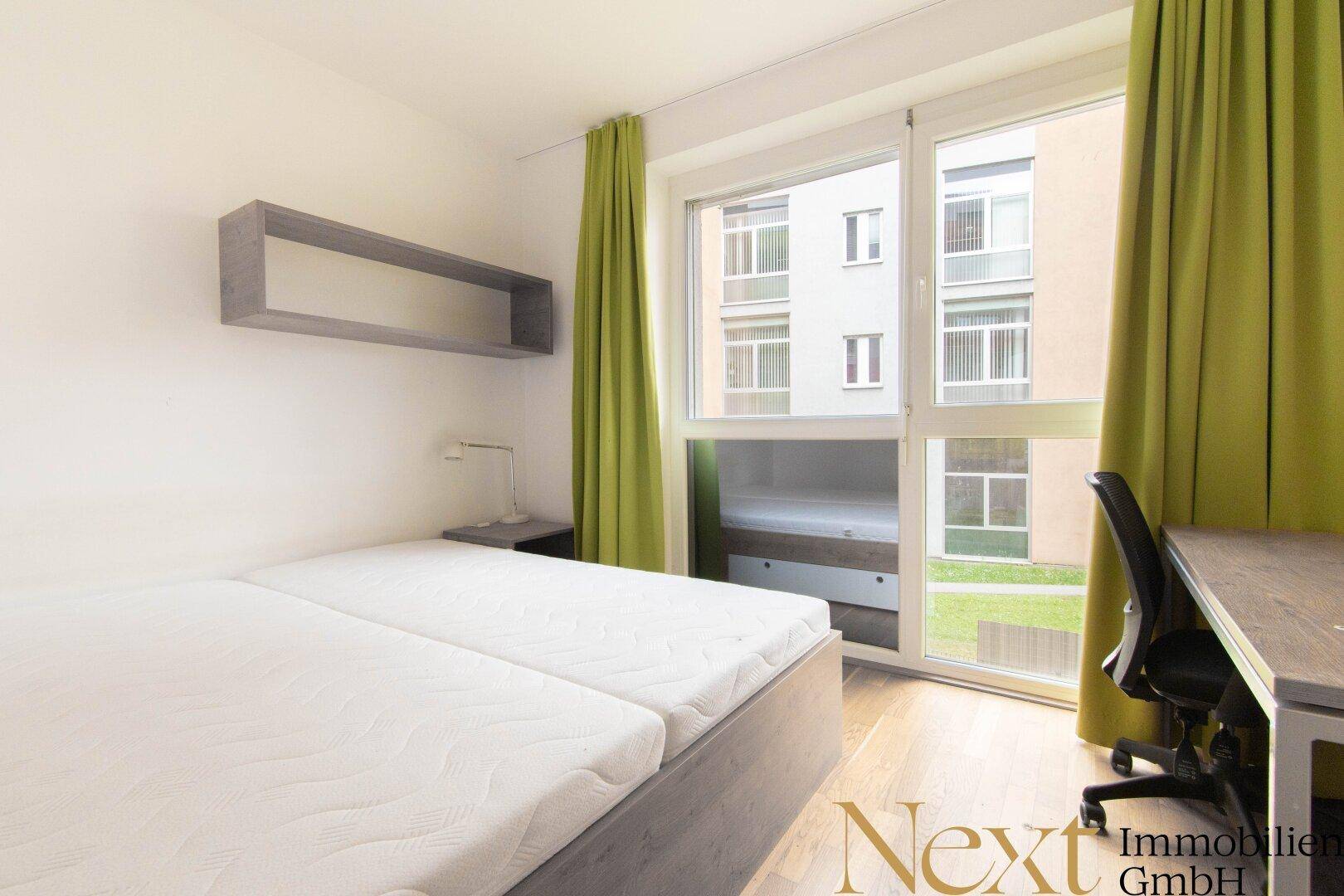 Funktionales Schlafzimmer mit zwei Einzelbetten, Schreibtisch und viel Tageslicht durch große Fenster.