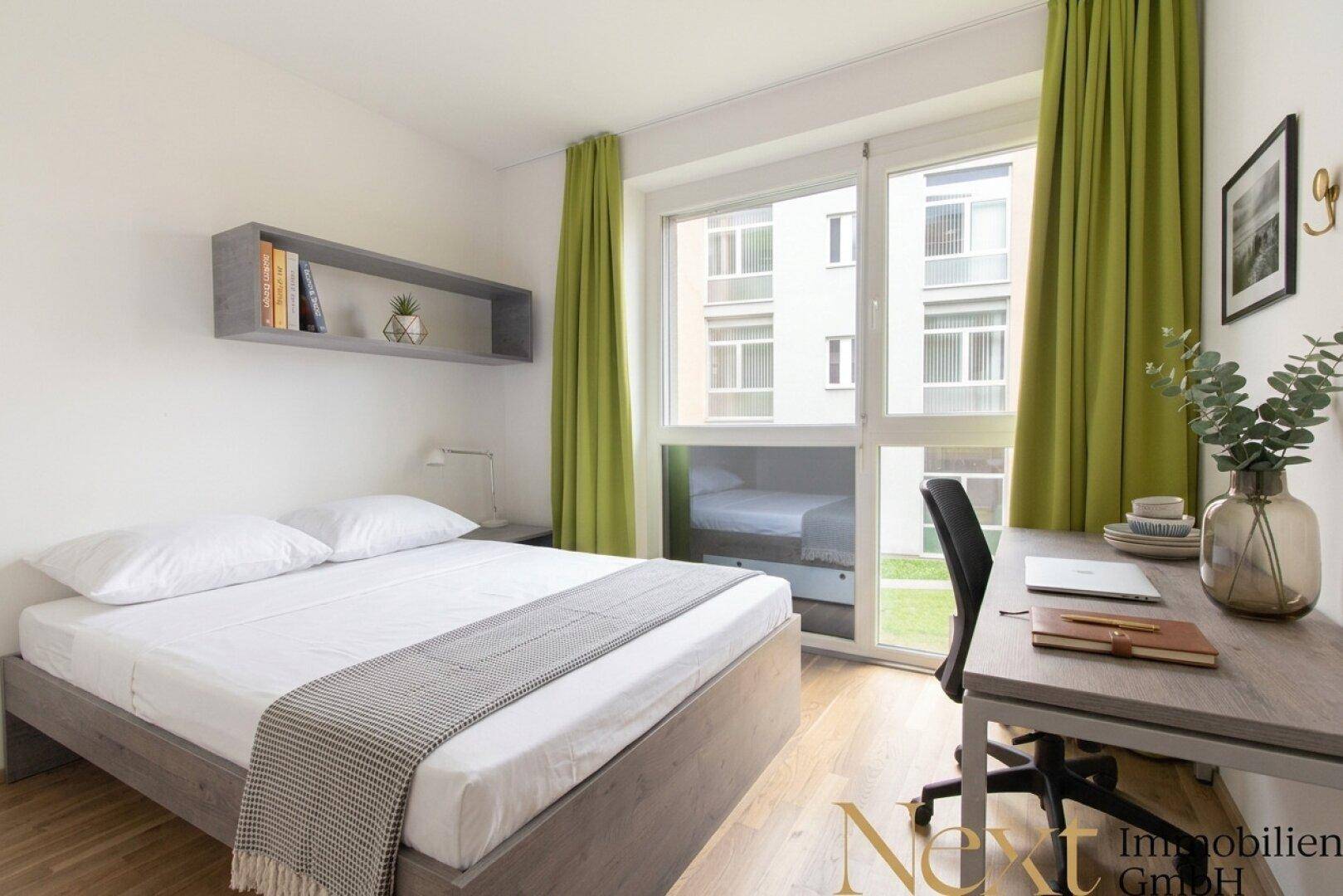 Modernes Schlafzimmer mit Einzelbett, Schreibtisch und großen Fenstern mit Blick auf den Innenhof.