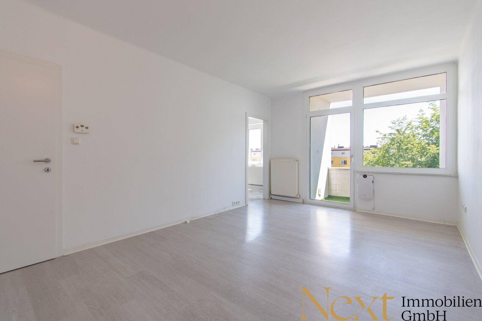 Geräumiges, leeres Wohnzimmer mit großen Fenstern und Zugang zu einem kleinen Balkon.