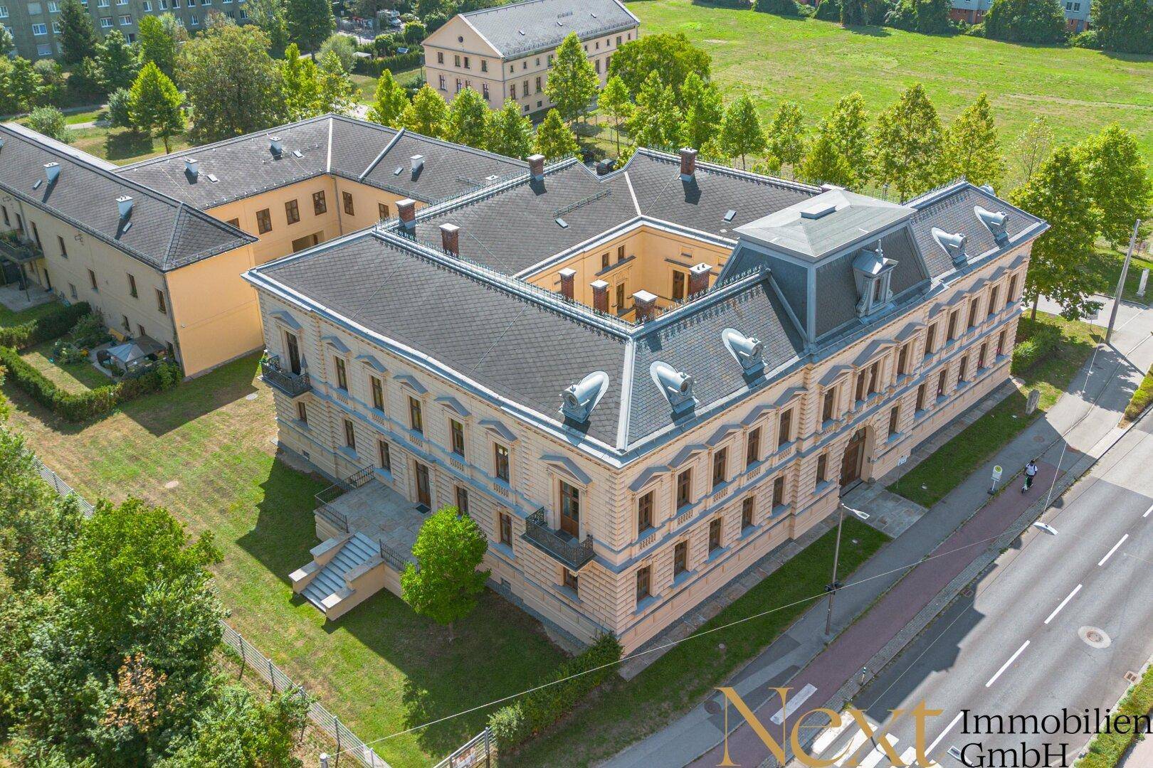 Luftaufnahme eines historischen Gebäudekomplexes mit Innenhof und umliegenden Grünflächen unter blauem Himmel.