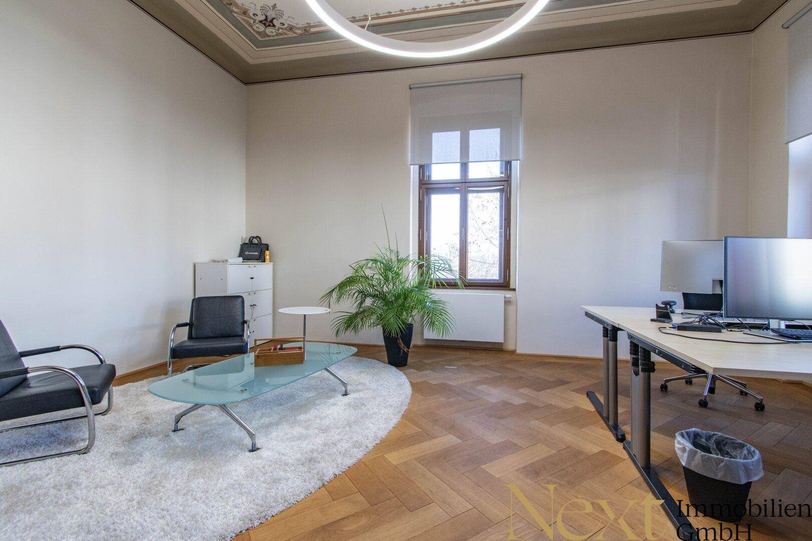 Helles Büro mit hohen Decken, Fischgrätparkett und moderner Einrichtung, durch ein großes Fenster natürlich belichtet.
