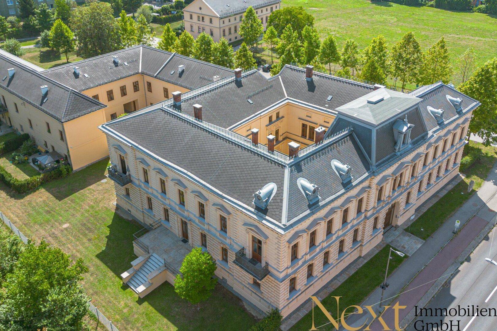 Luftaufnahme eines historischen Gebäudekomplexes mit Innenhof und umliegender Grünfläche unter blauem Himmel.