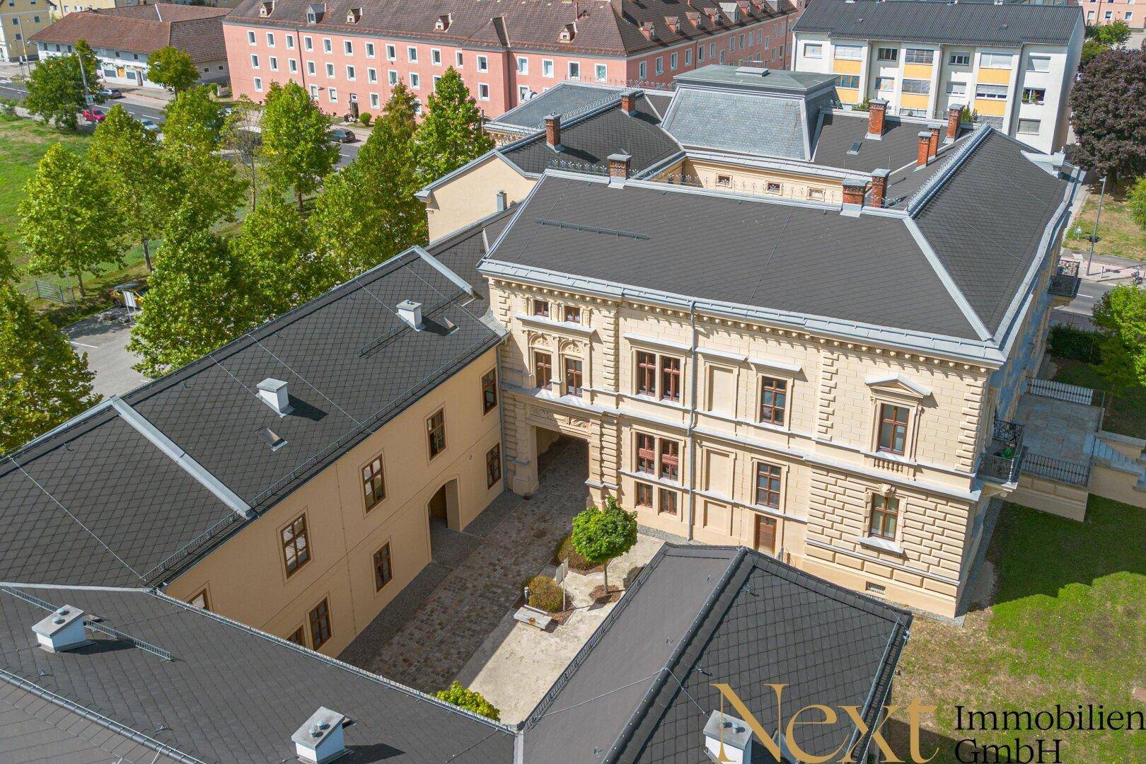 Blick von oben auf den weitläufigen Innenhof eines historischen Gebäudes mit gepflastertem Bereich und Bäumen.