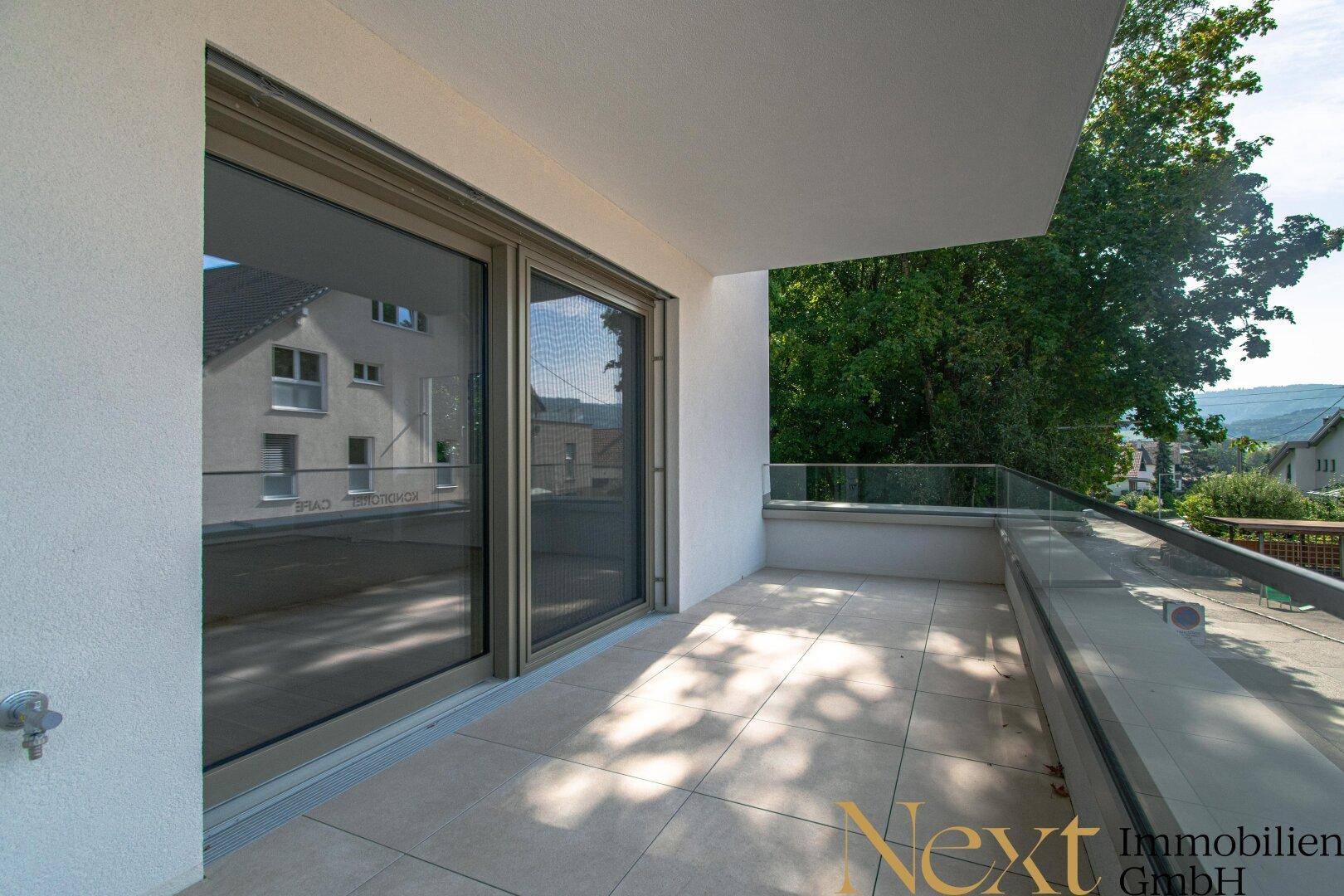 Geräumiger Balkon mit Fliesenboden und Glasgeländer, umgeben von grünen Bäumen.