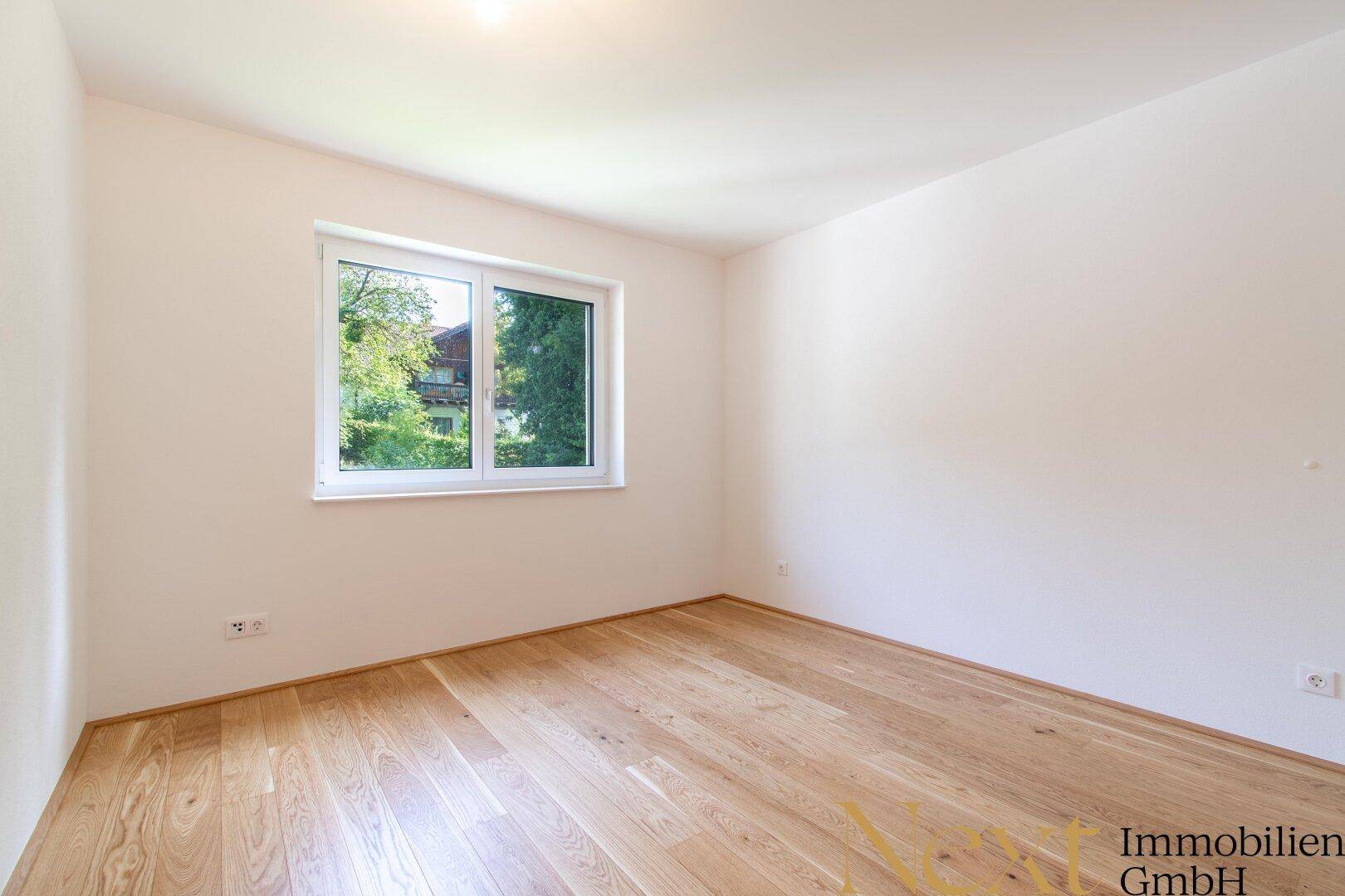 Gemütliches, leeres Schlafzimmer mit Holzboden und einem Fenster mit Blick ins Grüne.