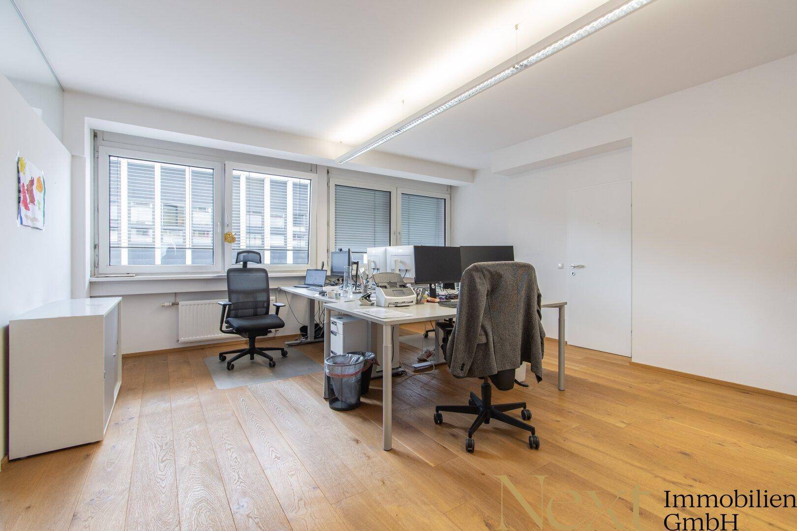 Helles Büro mit Holzboden und zwei Arbeitsplätzen, ausgestattet mit Computern und Bürostühlen.