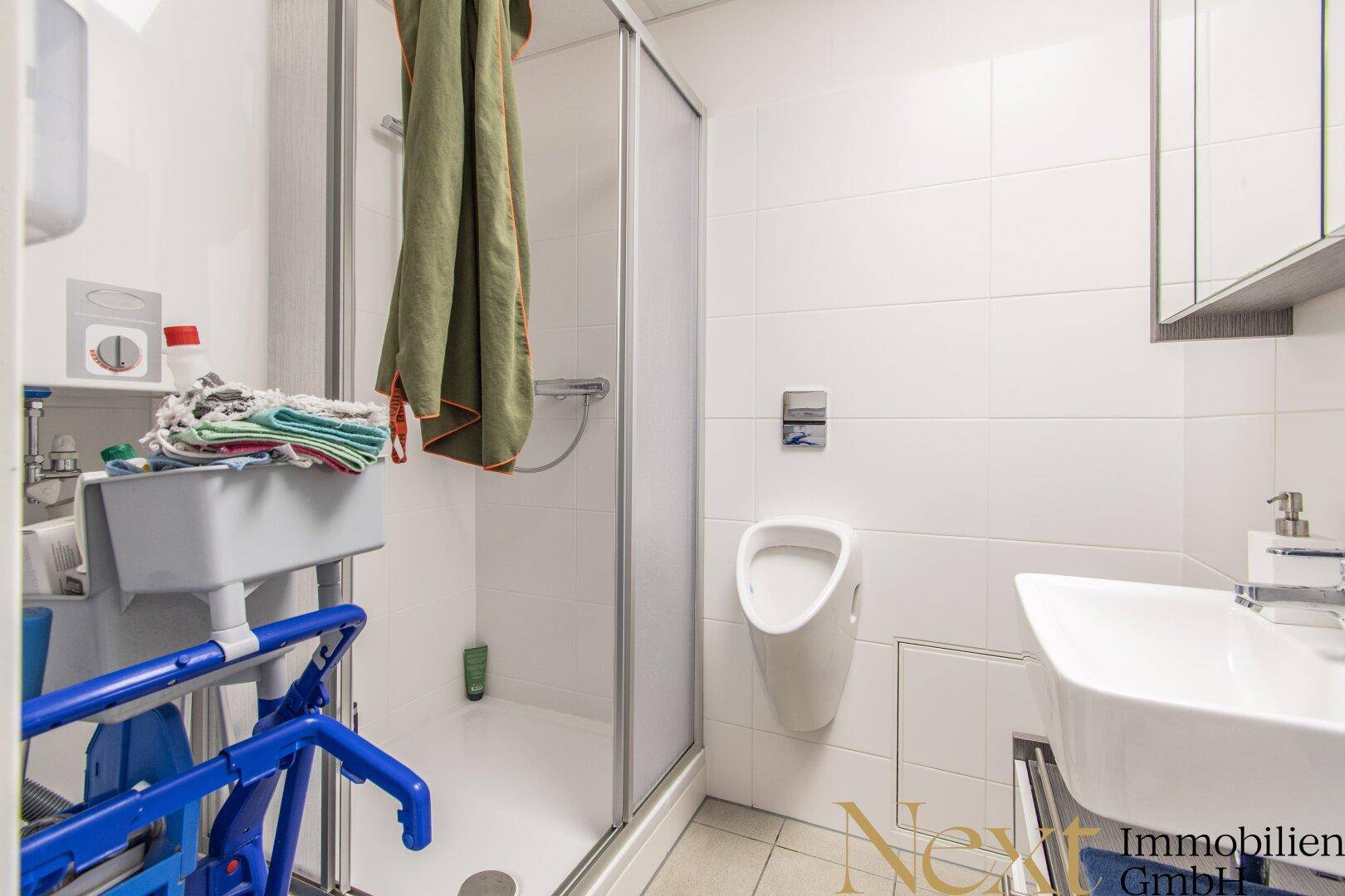 Modernes Badezimmer mit Dusche, Urinal und Waschbecken, alles in hellen Fliesen gehalten.
