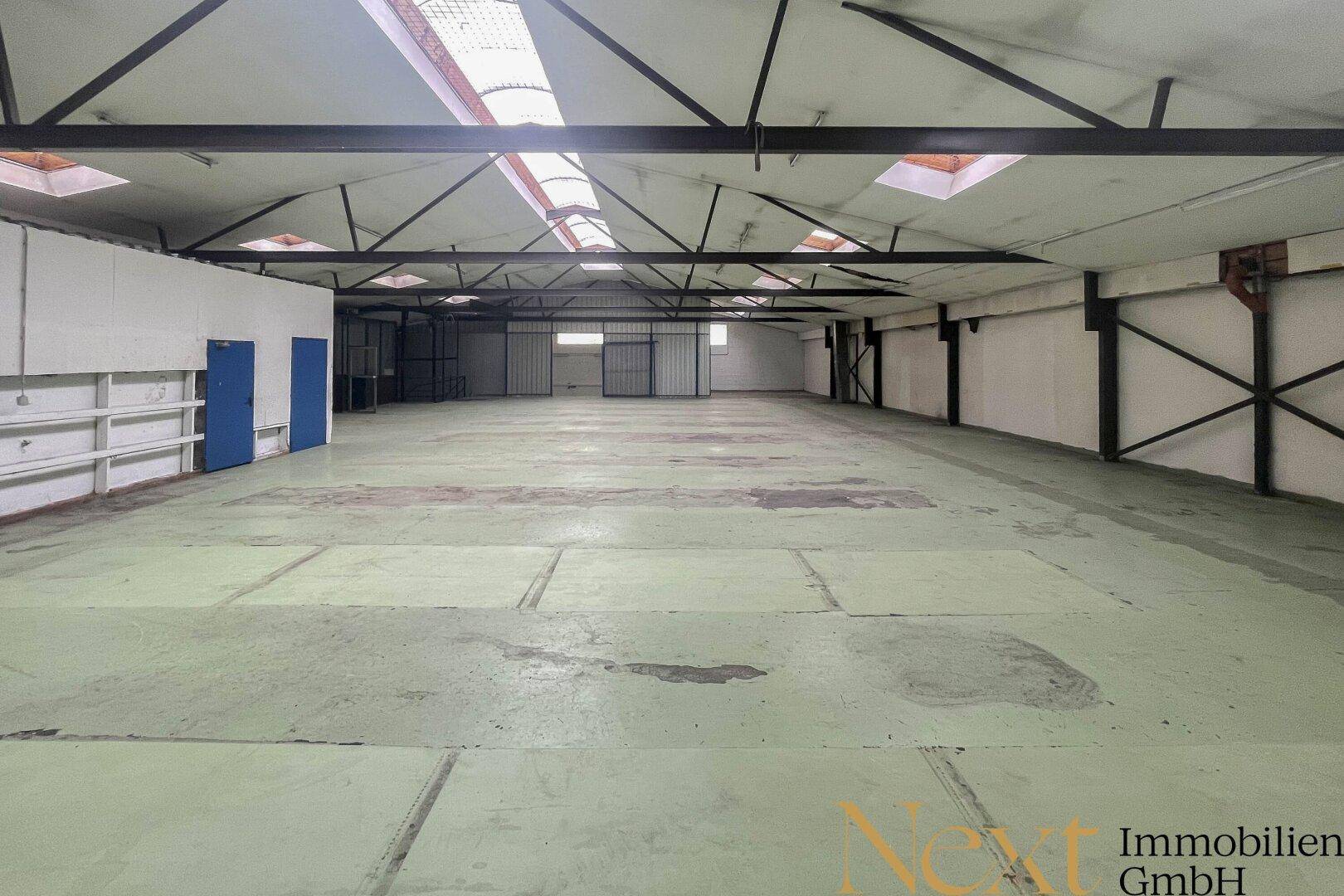 Große, leere Lagerhalle mit hohen Decken, Betonboden und Oberlichtern für natürliche Beleuchtung.