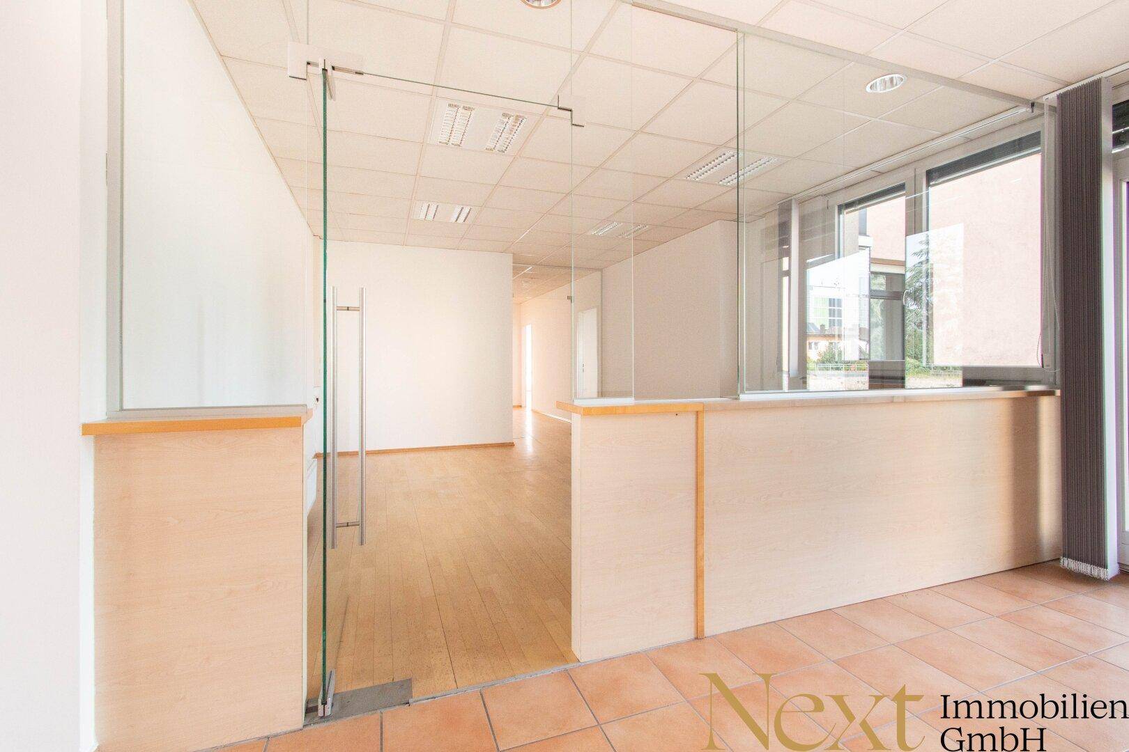 Bürobereich mit Glasabtrennungen und einer Glastür, die zu einem weiteren Raum mit Holzfußboden führt.