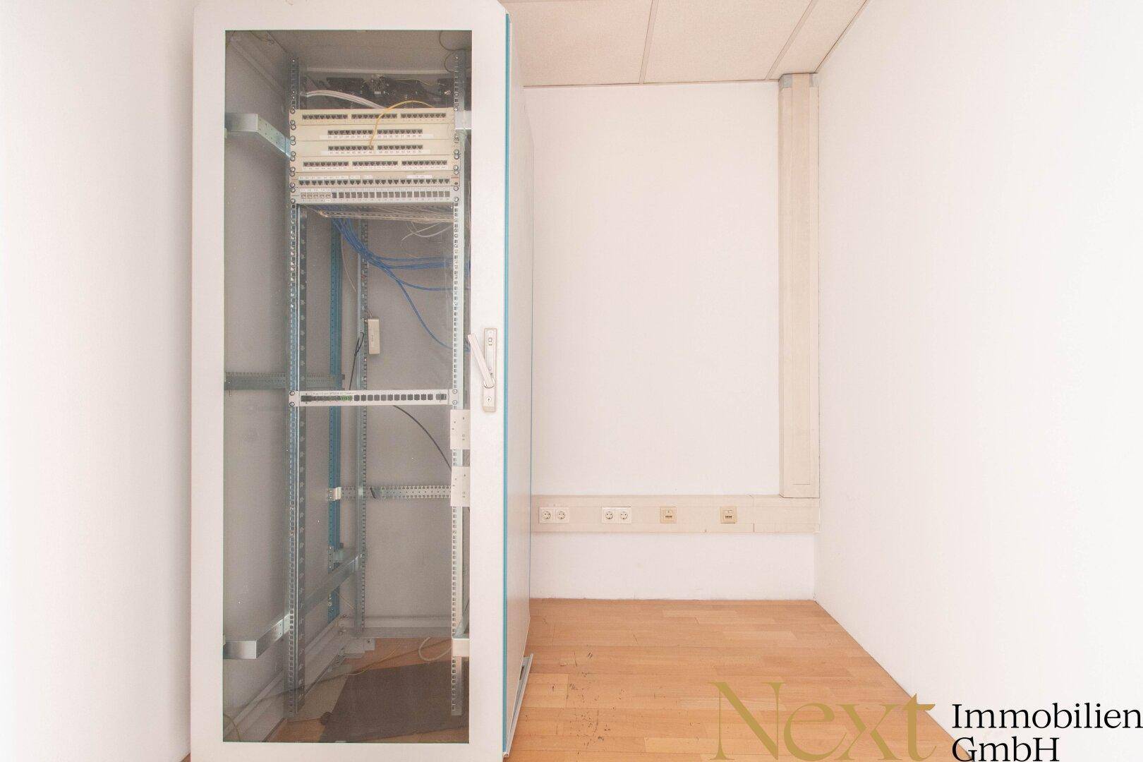 Kleiner Raum mit einem Serverschrank, Holzfußboden und zahlreichen Steckdosen für technische Geräte.