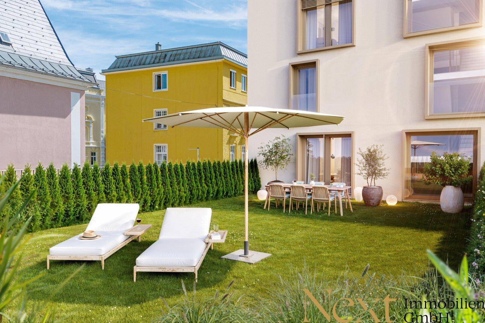 Sonniger Gartenbereich mit Liegestühlen, Esstisch und grüner Hecke vor moderner Fassade.