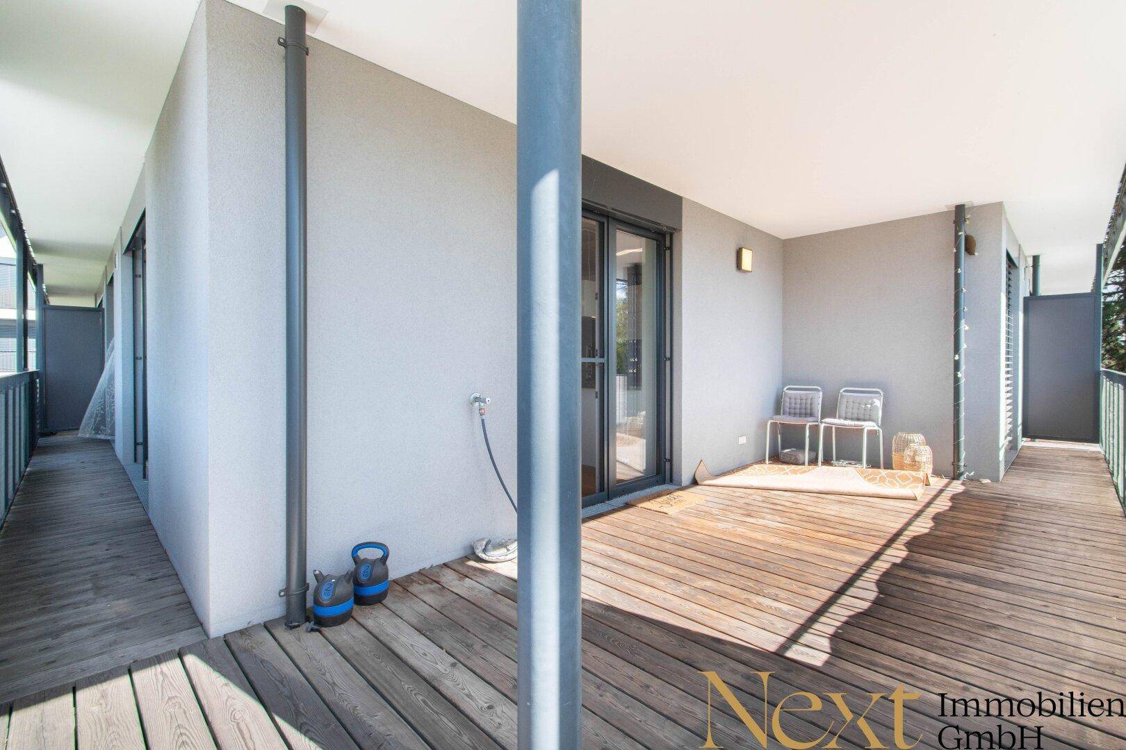 Langer Balkon mit Holzboden und Zugang zu mehreren Räumen, umgeben von Grün.