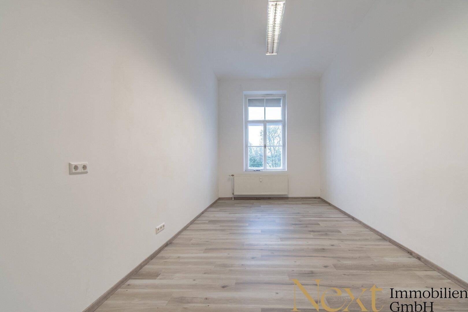 Leerer Raum mit hellen Wänden, Laminatboden und einem Fenster, das Tageslicht hereinlässt.