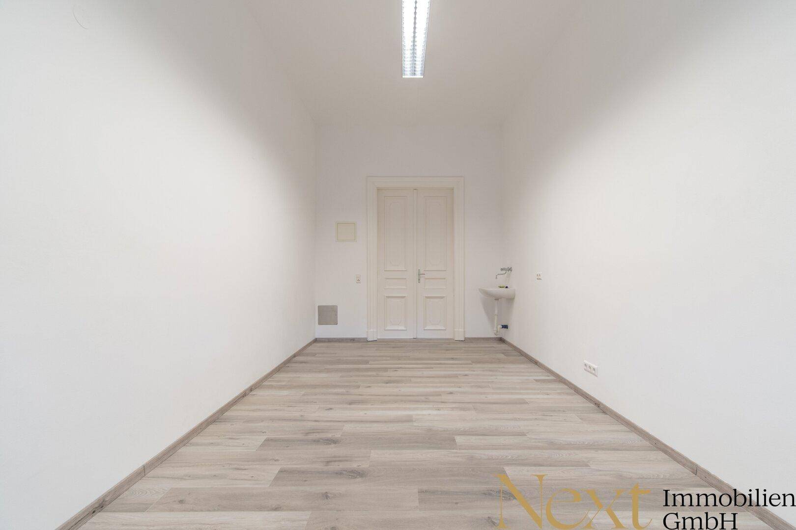 Leerer Raum mit weißen Wänden, Laminatboden und einer Doppeltür am Ende, inklusive kleinem Waschbecken.