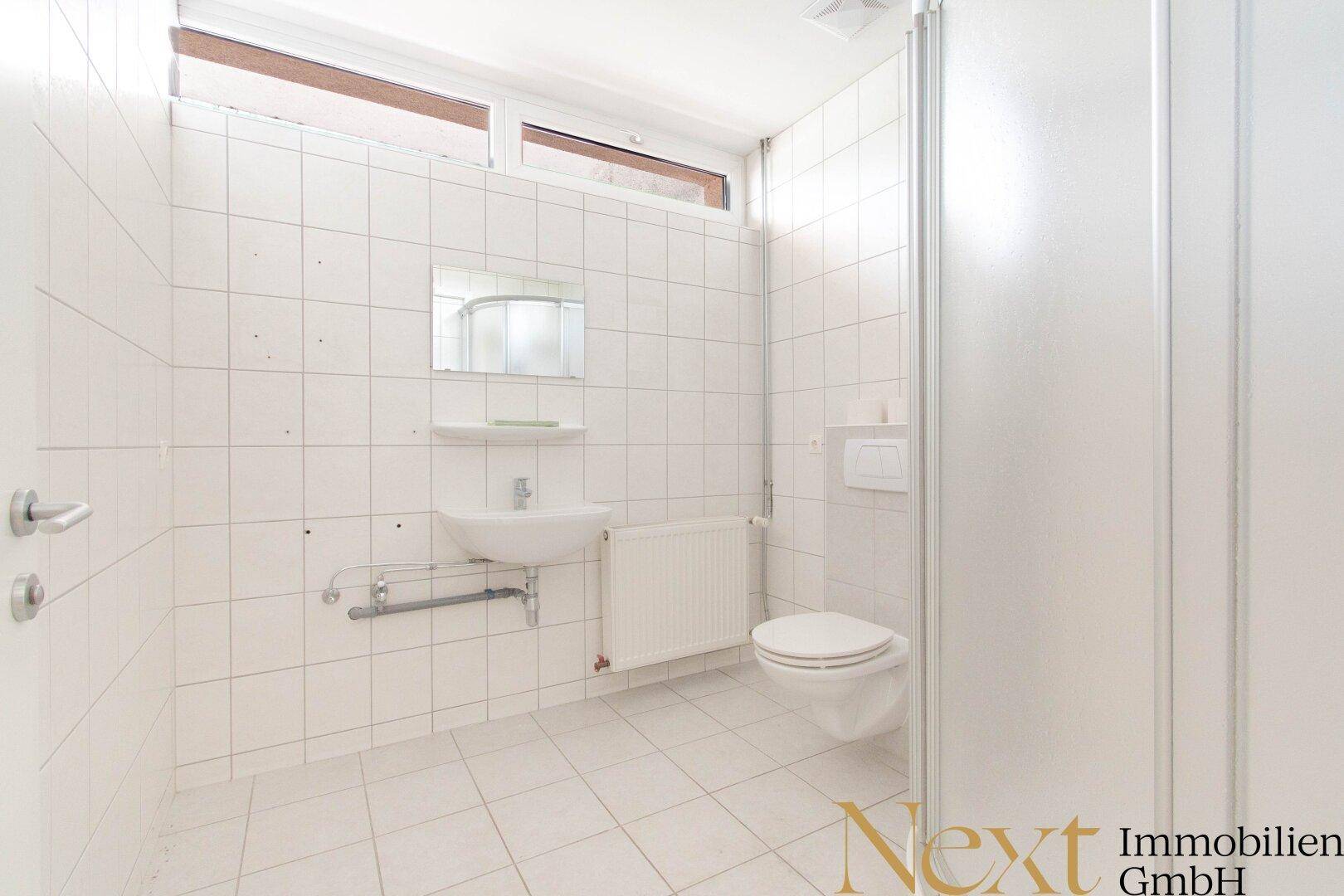 Funktionales Badezimmer mit weißen Fliesen, Dusche, WC und einem kleinen Fenster für Belüftung.