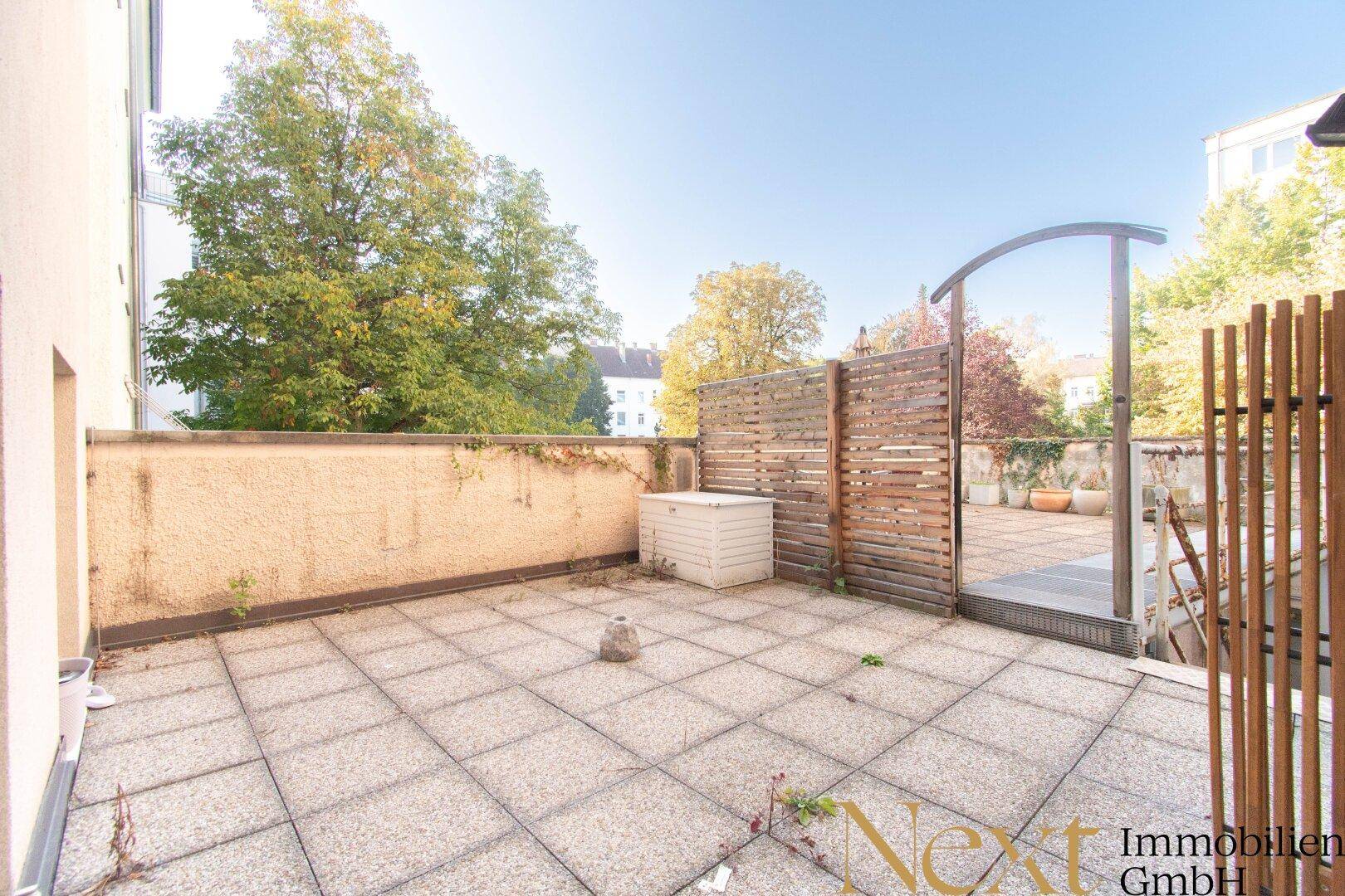 Geräumige Terrasse mit Steinplatten und Holzelementen, umgeben von Bäumen und Grünflächen.