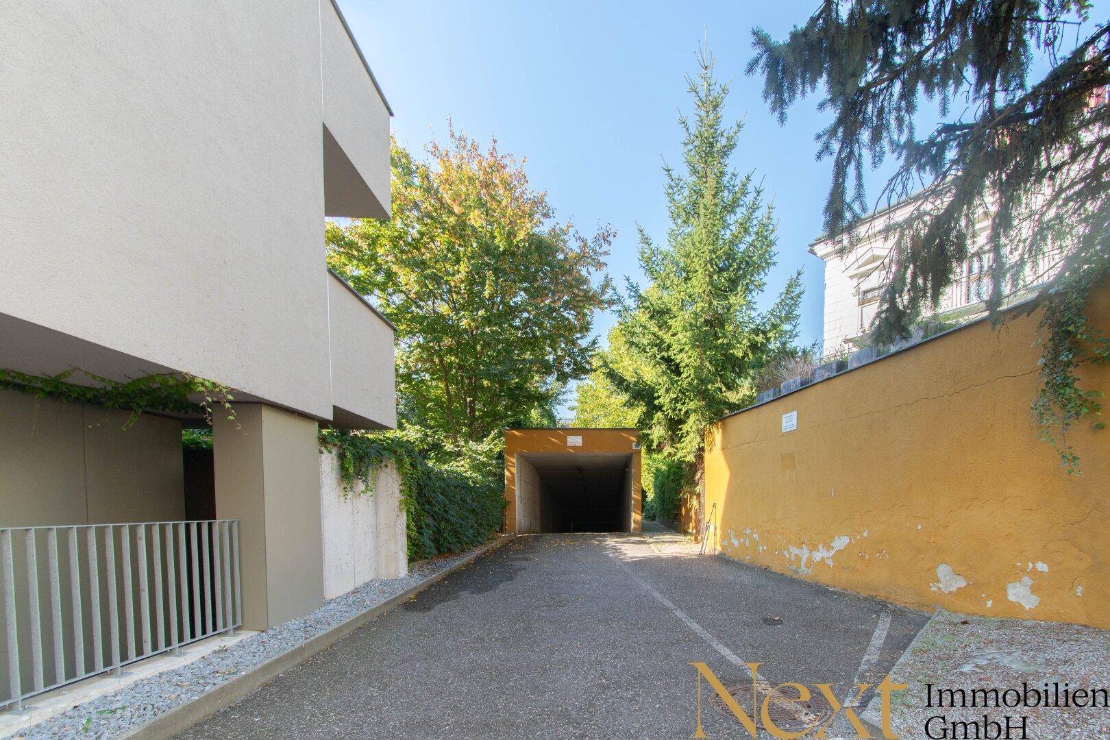 Einfahrt zur Tiefgarage, umgeben von Bäumen und Gebäuden mit einer gelben Fassade.