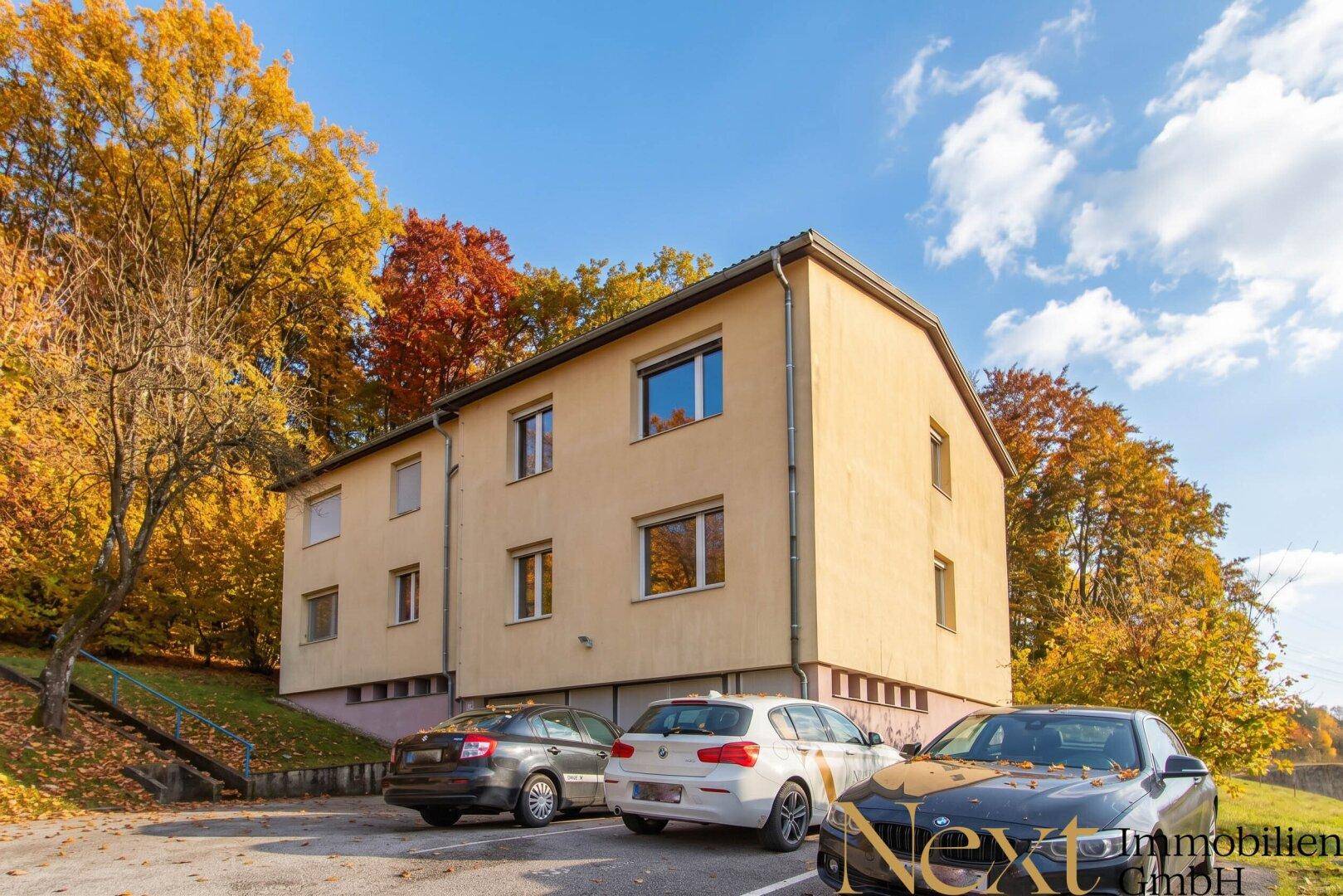 Mehrfamilienhaus mit Parkplätzen und umgebender Natur im Herbst.