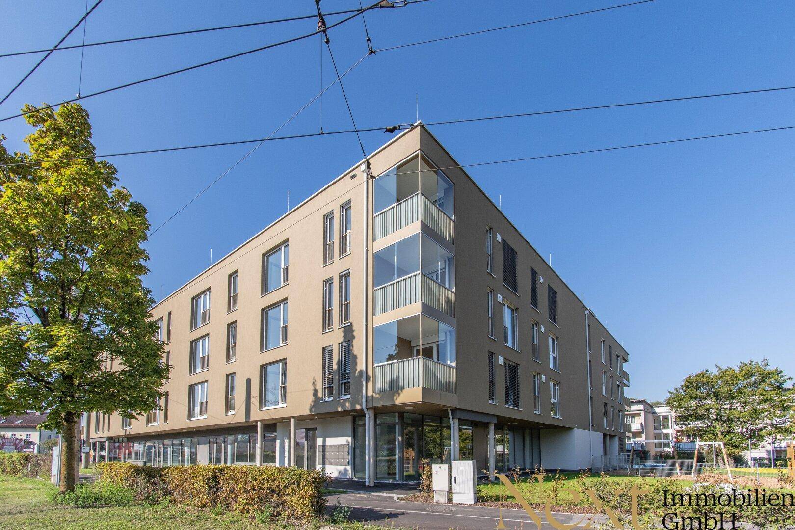 Modernes Mehrfamilienhaus mit beiger Fassade und großen Fenstern in städtischer Umgebung unter blauem Himmel.