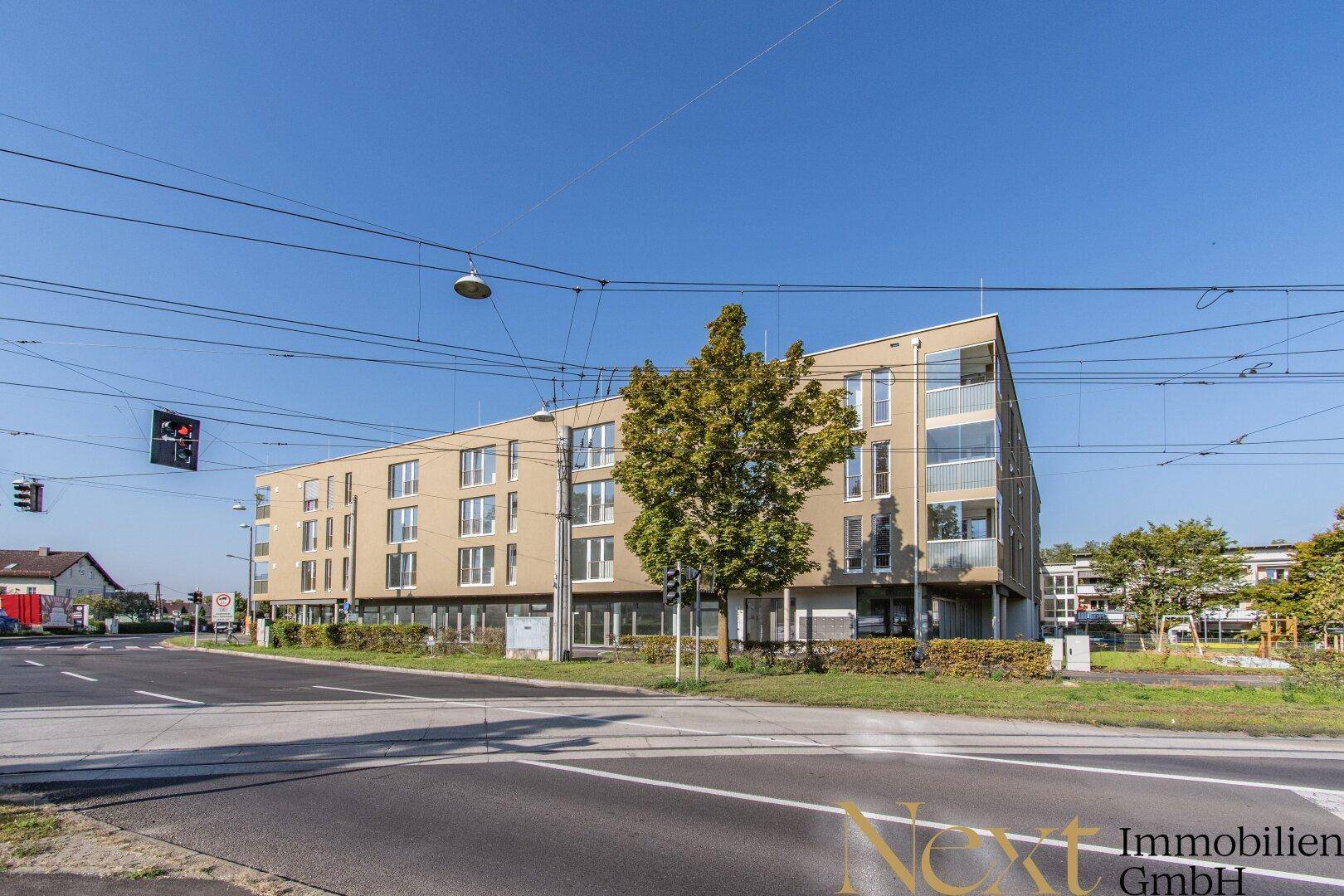 Neubau-Gebäude an einer belebten Straße mit Straßenbahnschienen und klarem Himmel, umgeben von Grünflächen.