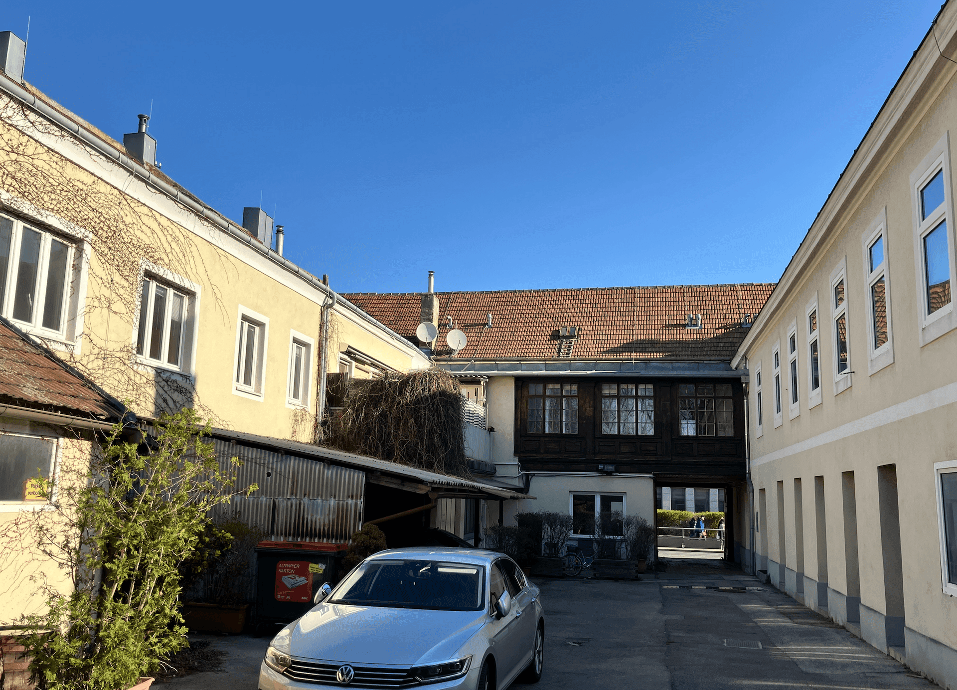 Blick in einen Innenhof mit mehreren Gebäuden, einem geparkten Auto und klarem blauem Himmel.