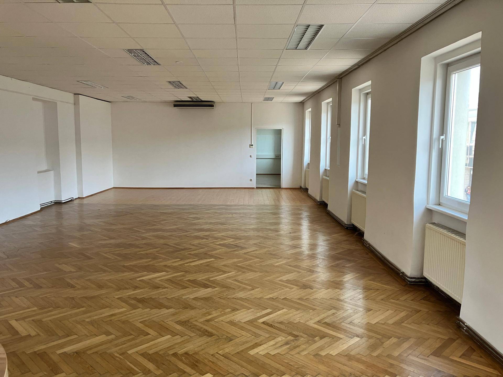 Geräumiger, leerer Raum mit glänzendem Fischgrätparkett, weißen Wänden und viel Tageslicht durch Fenster.