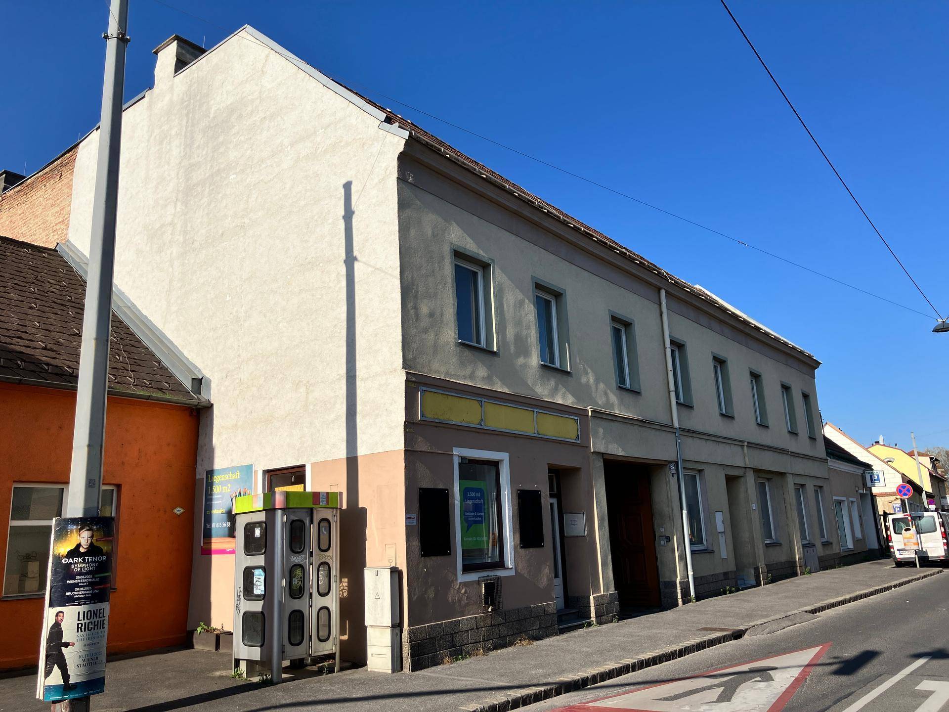 Straßenansicht eines Gebäudes mit hellen Fassaden, Geschäftsfront im Erdgeschoss und einem Telefonhäuschen.