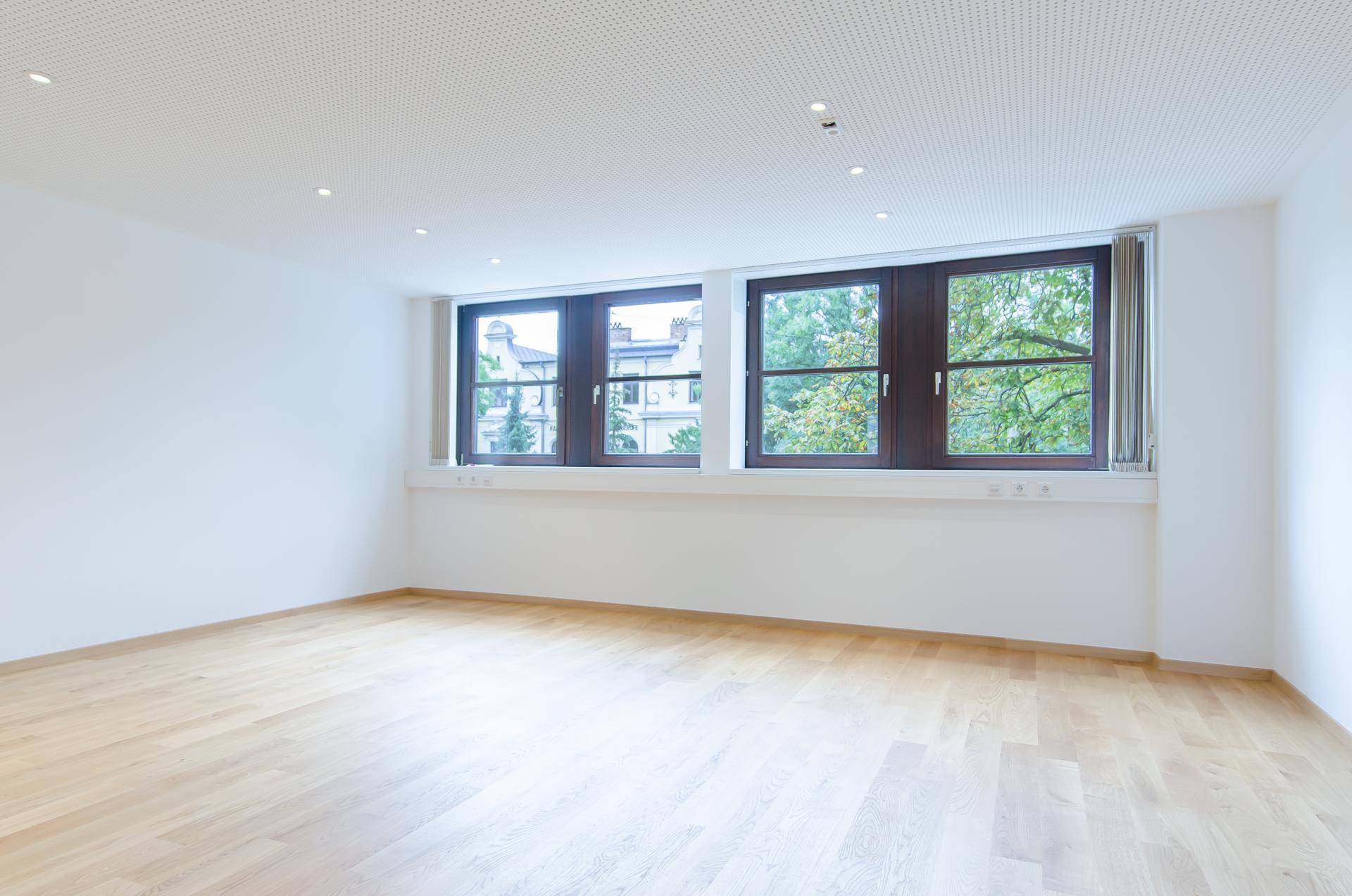 Ein leerer Büroraum mit hellem Holzparkett und großen Fenstern, die viel Tageslicht hereinlassen.