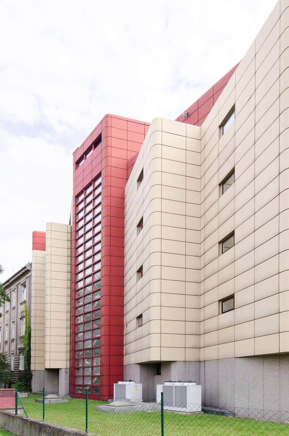 Moderne Gebäudefassade mit markanten roten und beigen Paneelen sowie einer vertikalen Glasfront, umgeben von Grünflächen.