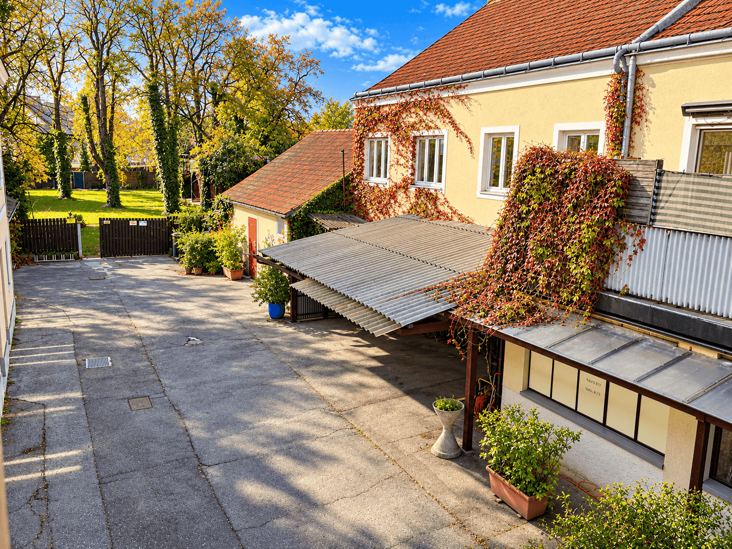 Innenhof mit Gebäuden, üppiger Vegetation und einem überdachten Bereich unter blauem Himmel.