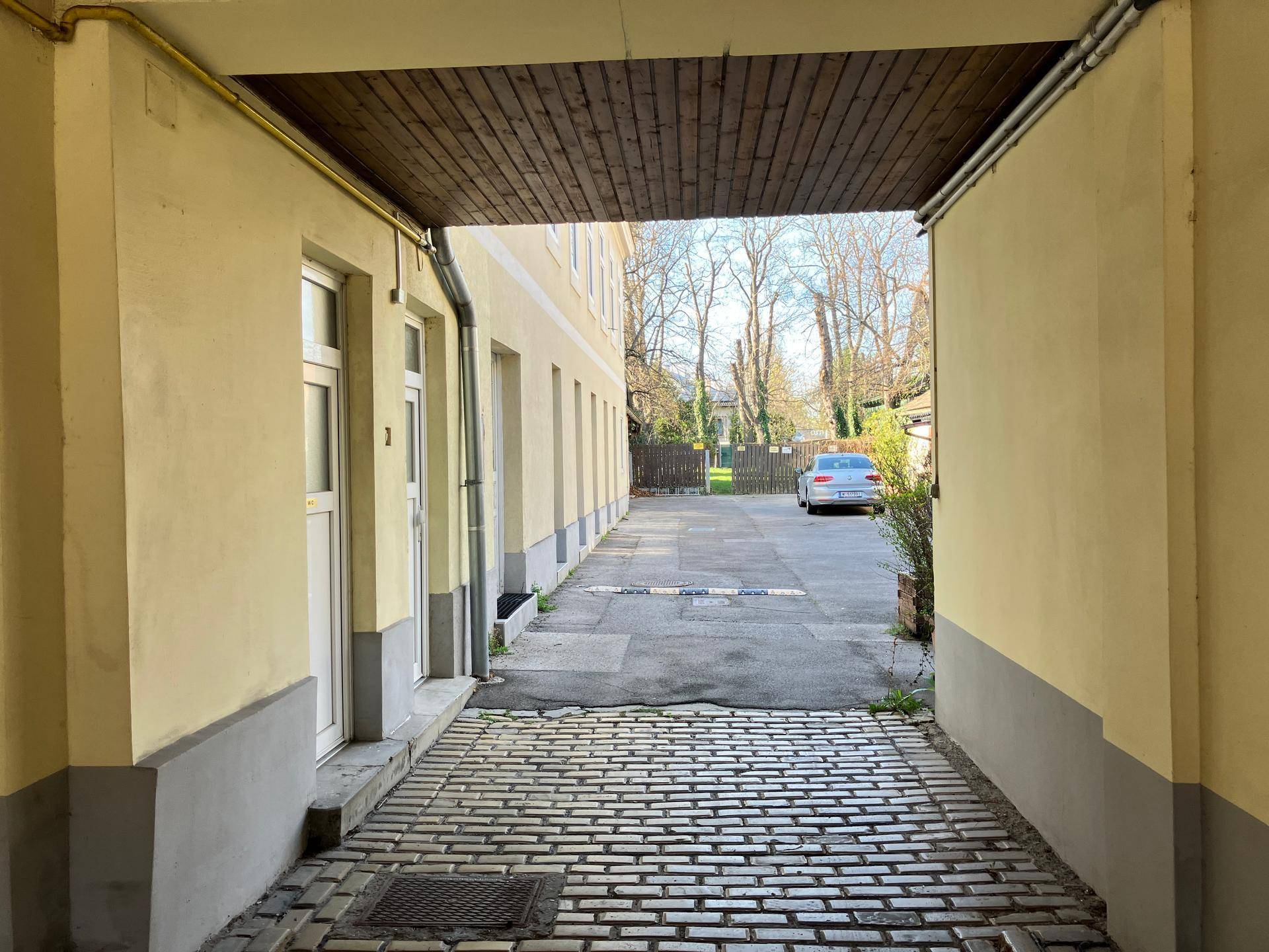 Durchgang mit Kopfsteinpflaster führt zu einem offenen Bereich mit geparktem Auto und Bäumen im Hintergrund.