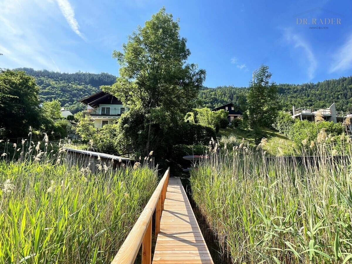 Langer Holzsteg durch Schilf mit Blick auf das Haus und die umliegende Natur.