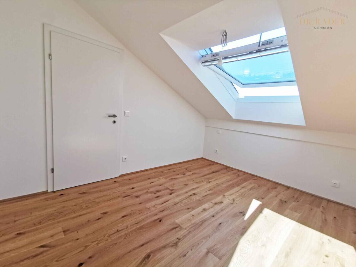 Leeres Zimmer im Dachgeschoss mit Holzboden, weiß gestrichenen Wänden und einem Dachfenster.