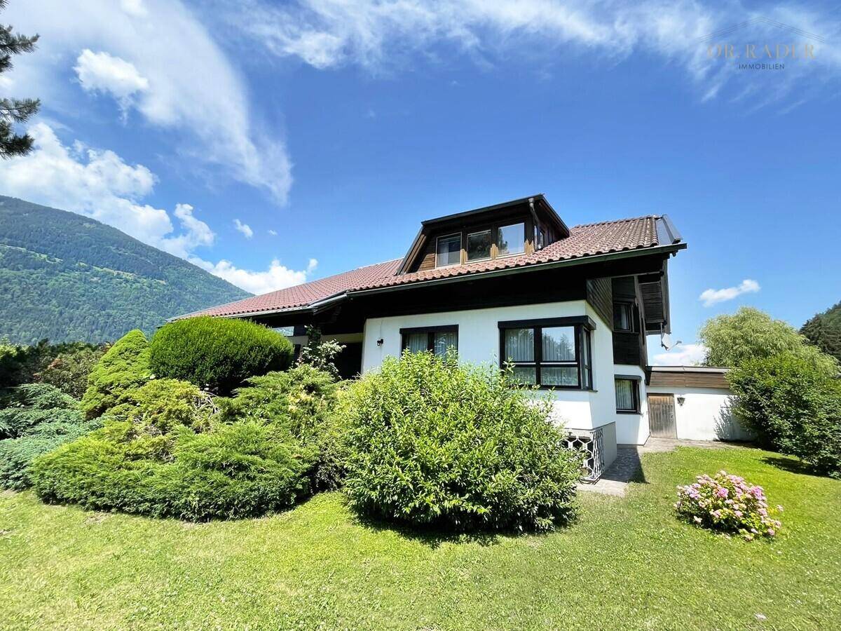 Charmantes Haus mit weißer Fassade, dunklem Dach und gepflegtem Vorgarten vor Bergpanorama.