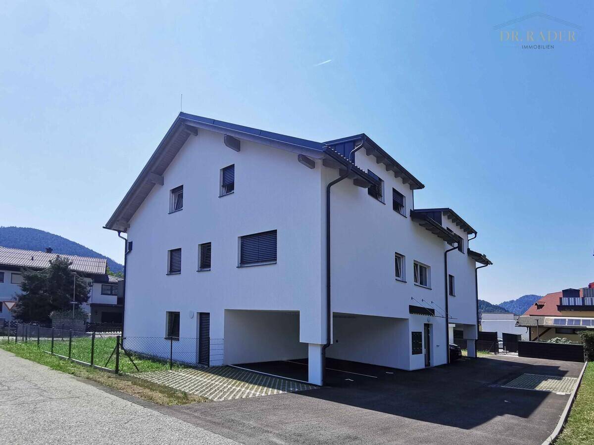 Rückansicht des Neubaus mit integrierten Carports und weißer Fassade vor einer grünen Hügellandschaft.