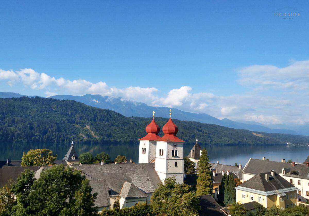 Malerische Stadtansicht mit See, Bergen und einer Kirche mit roten Zwiebeltürmen unter blauem Himmel.