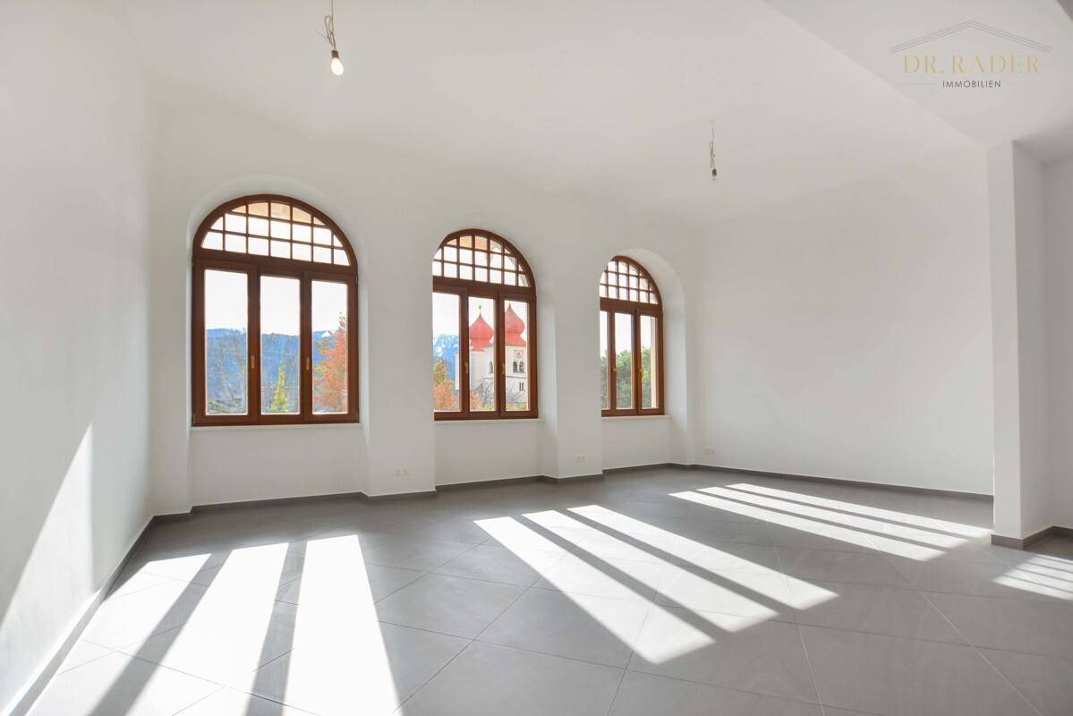 Heller, leerer Raum mit großen Rundbogenfenstern, die viel Sonnenlicht hereinlassen und einen Blick auf die Berge bieten.