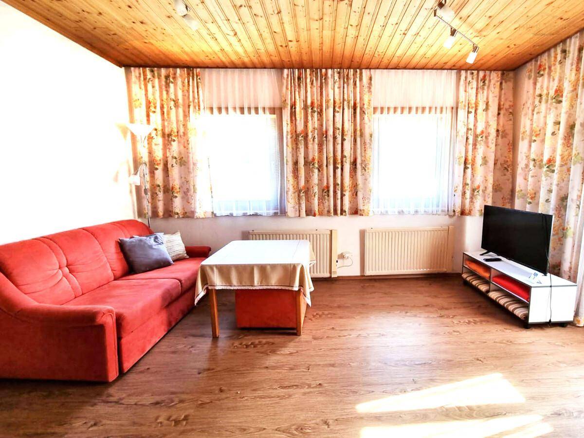 Geräumiges Wohnzimmer mit rotem Sofa, Holzboden und zwei Fenstern.