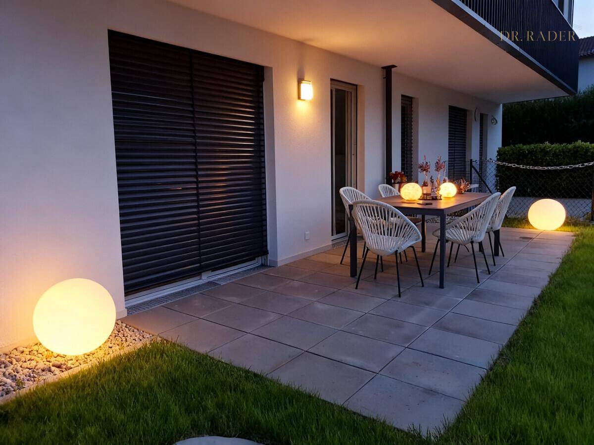 Gemütliche Terrasse am Abend mit Esstisch, Stühlen und stimmungsvoller Beleuchtung neben einer Rasenfläche.
